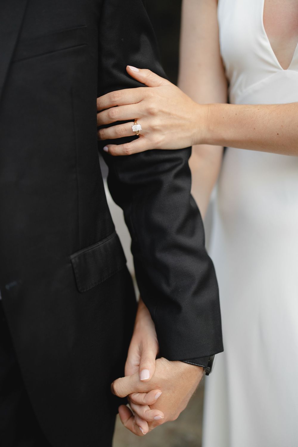 Close-up of bride's engagement ring as she holds groom's arm and hand in black suit sleeve.