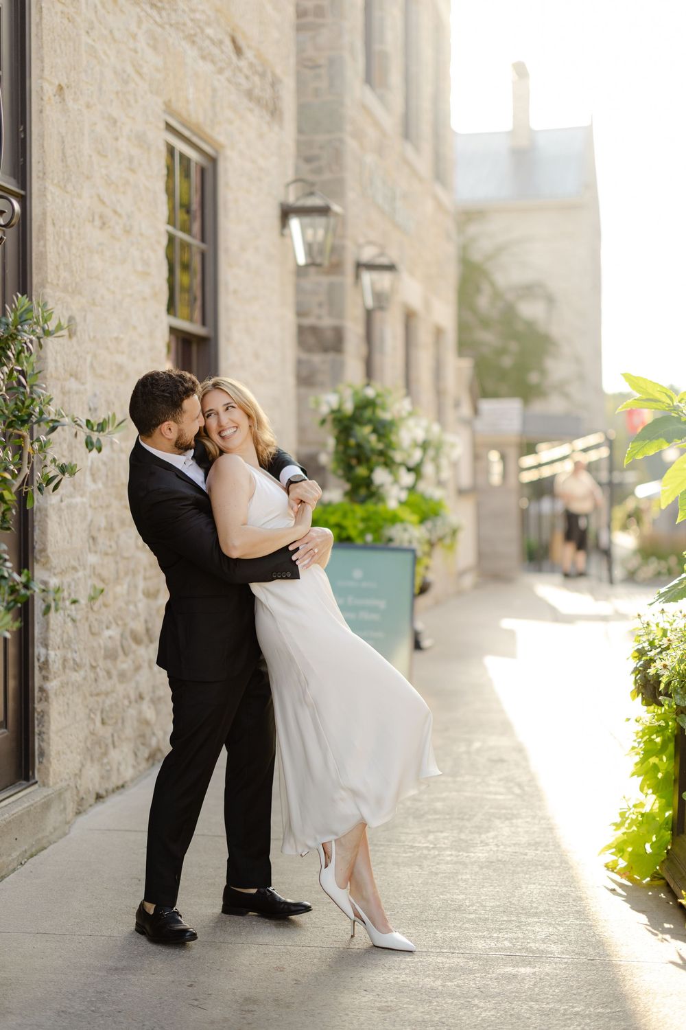 Bride and groom embracing and laughing outside a stone building on their wedding day.