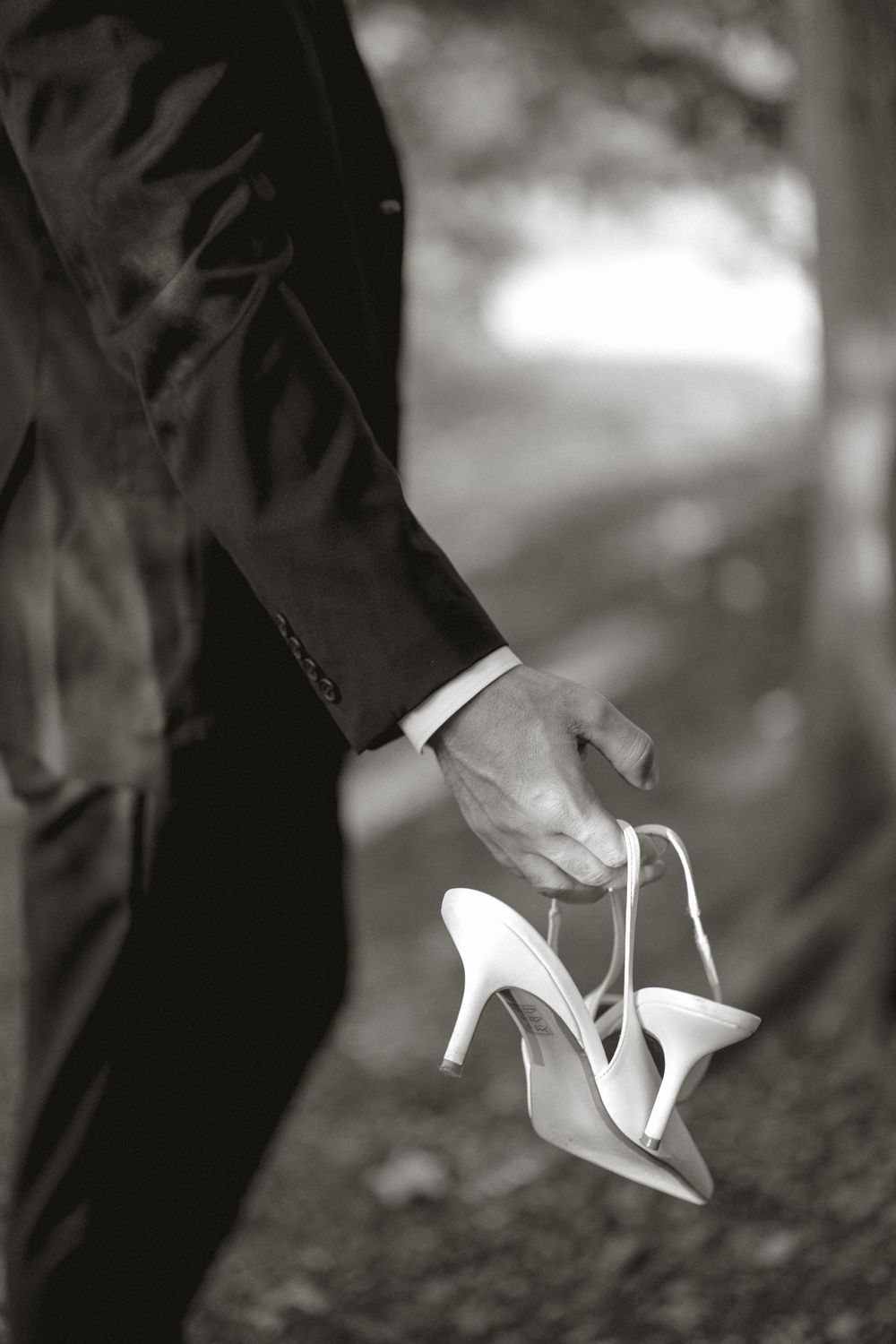 Black and white photo of groom carrying brides white high heel shoes in a forest.
