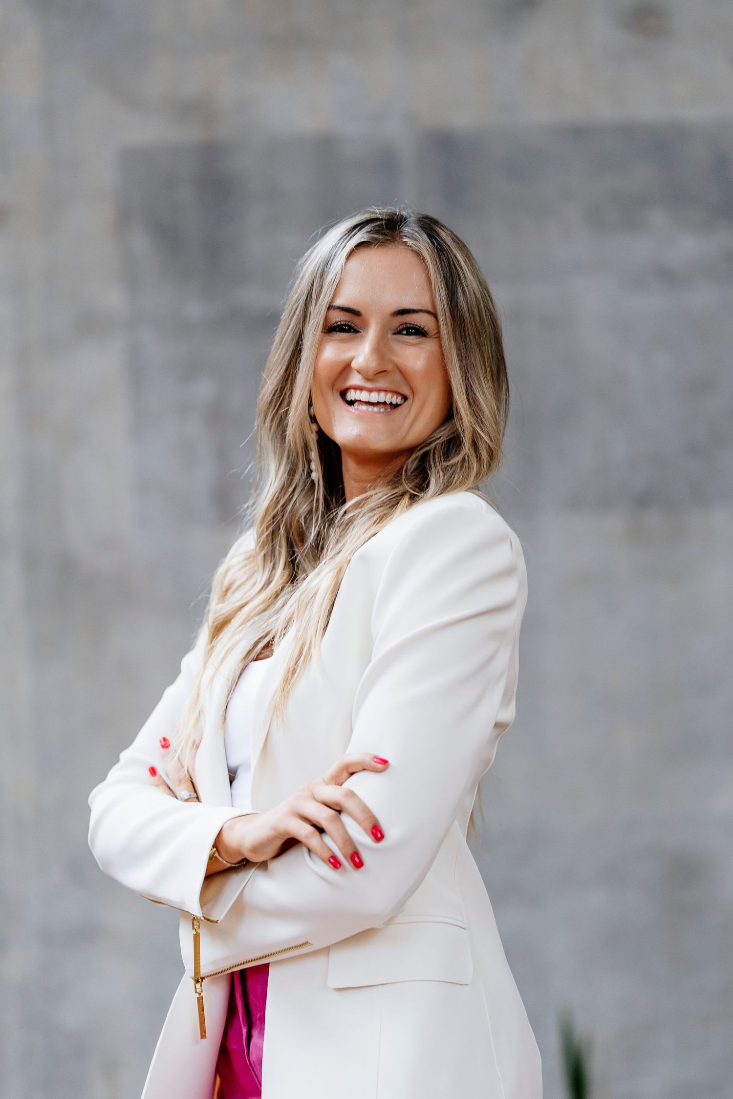 Smiling blonde woman in white blazer with arms crossed, posing confidently against a gray concrete wall headshot photographer queensland
