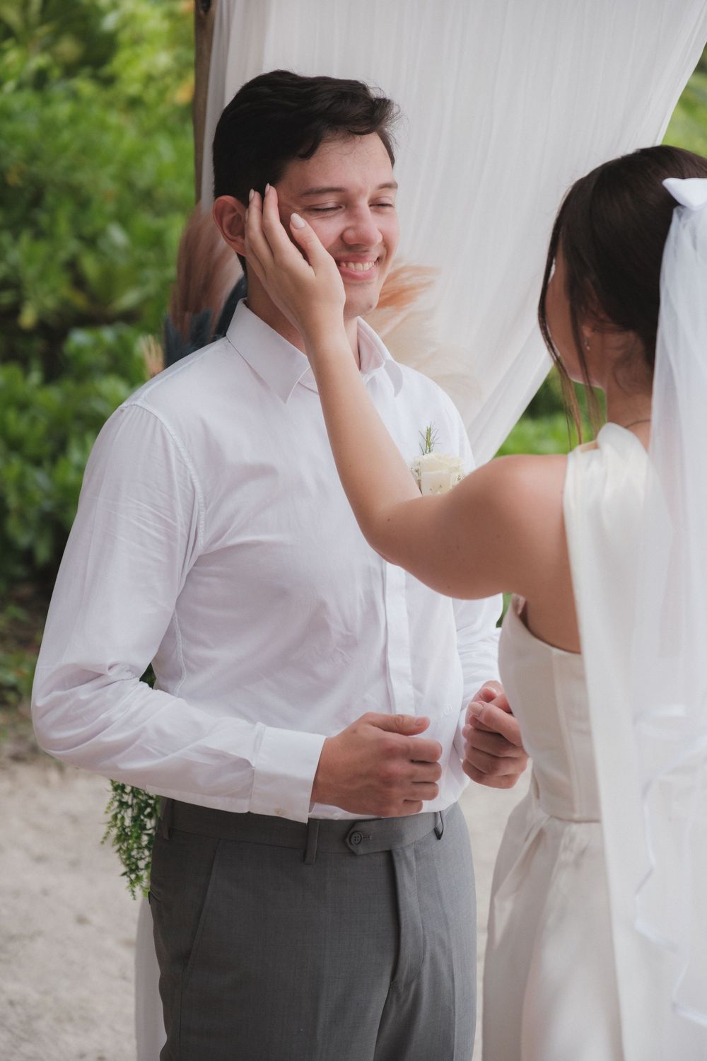 Groom smiling as bride tenderly touches his face during an outdoor beach wedding ceremony.