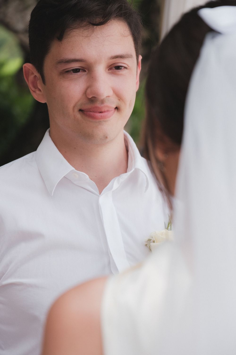 Groom in white shirt with boutonniere smiling at bride in veil during outdoor wedding ceremony.
