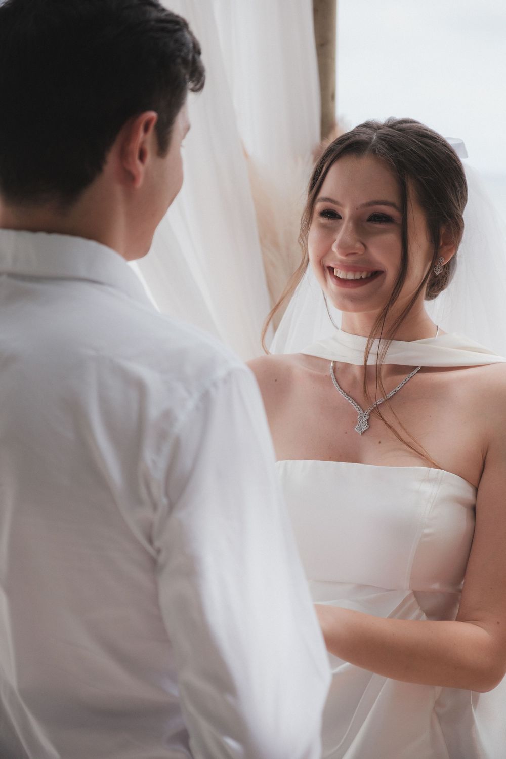 Smiling bride in white strapless gown and veil shares a joyful moment with groom near a sunlit window.