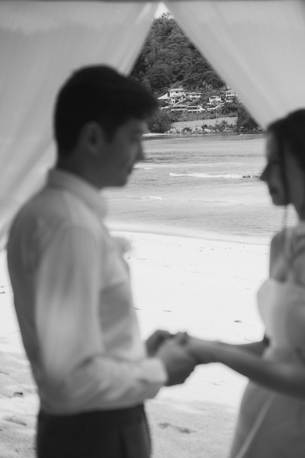 Black and white beach wedding ceremony with couple exchanging rings under draped canopy with ocean and tropical hillside backdrop.