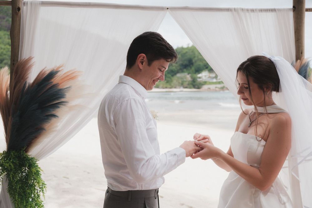 A groom places a wedding ring on his bride's finger during a romantic beach ceremony under a white draped canopy.