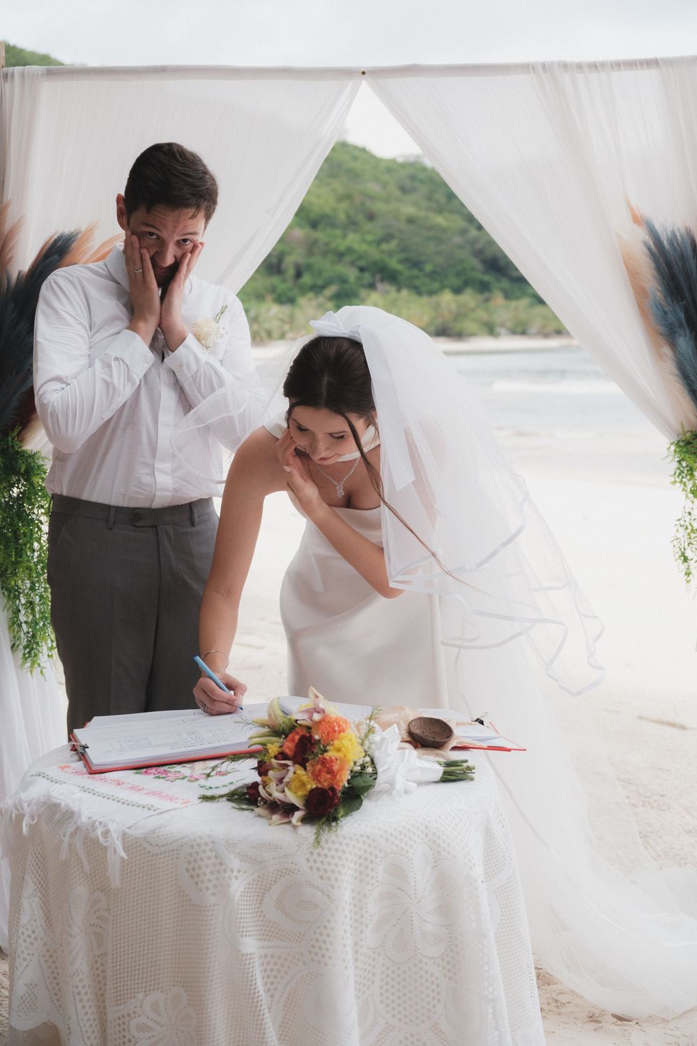 Bride and groom signing wedding registry at beach ceremony altar with white drapes and tropical greenery.