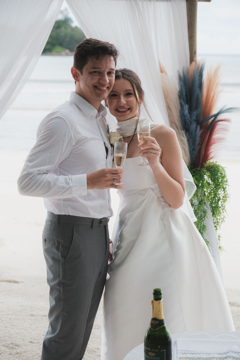 Newlywed couple toasting with champagne glasses on a beach wedding venue with white drapes.