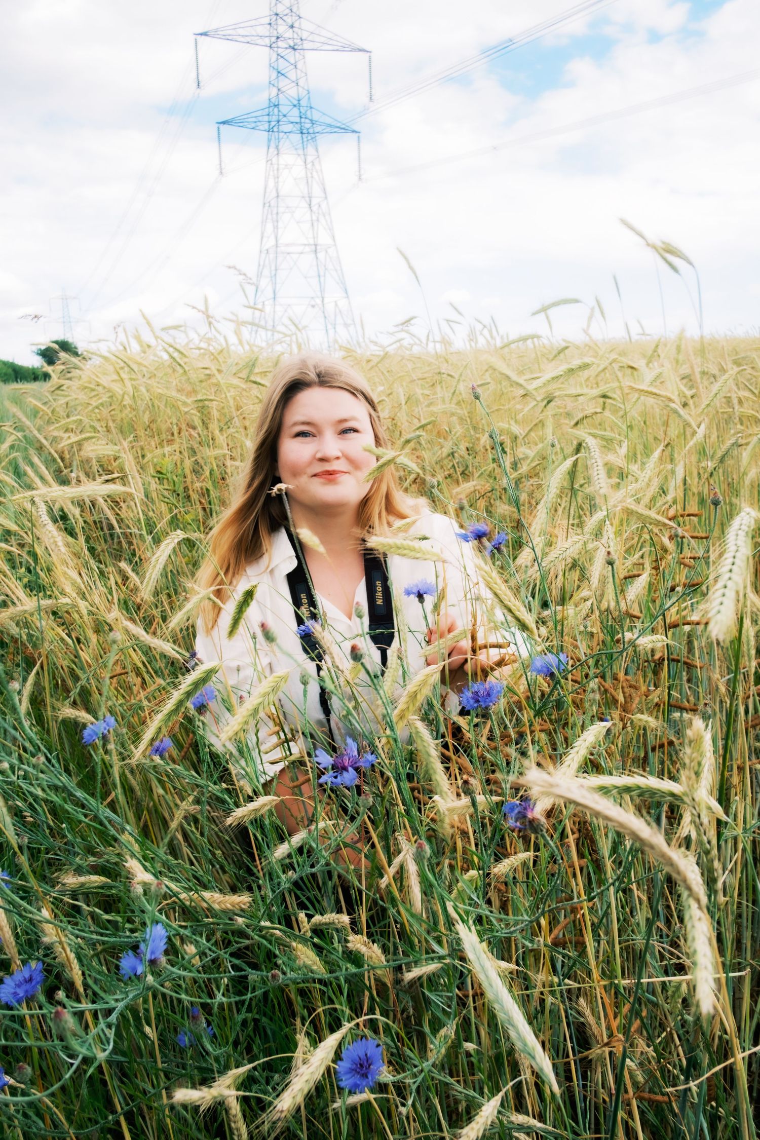 Suffolk Wildlife Staff: Woman smiling in a golden wheat field with blue cornflowers and an electricity pylon in the background.