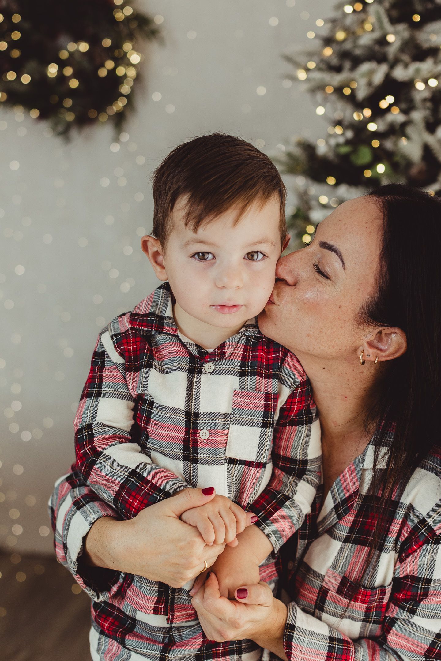 Mother and toddler in matching plaid pajamas share a kiss by a decorated Christmas tree.