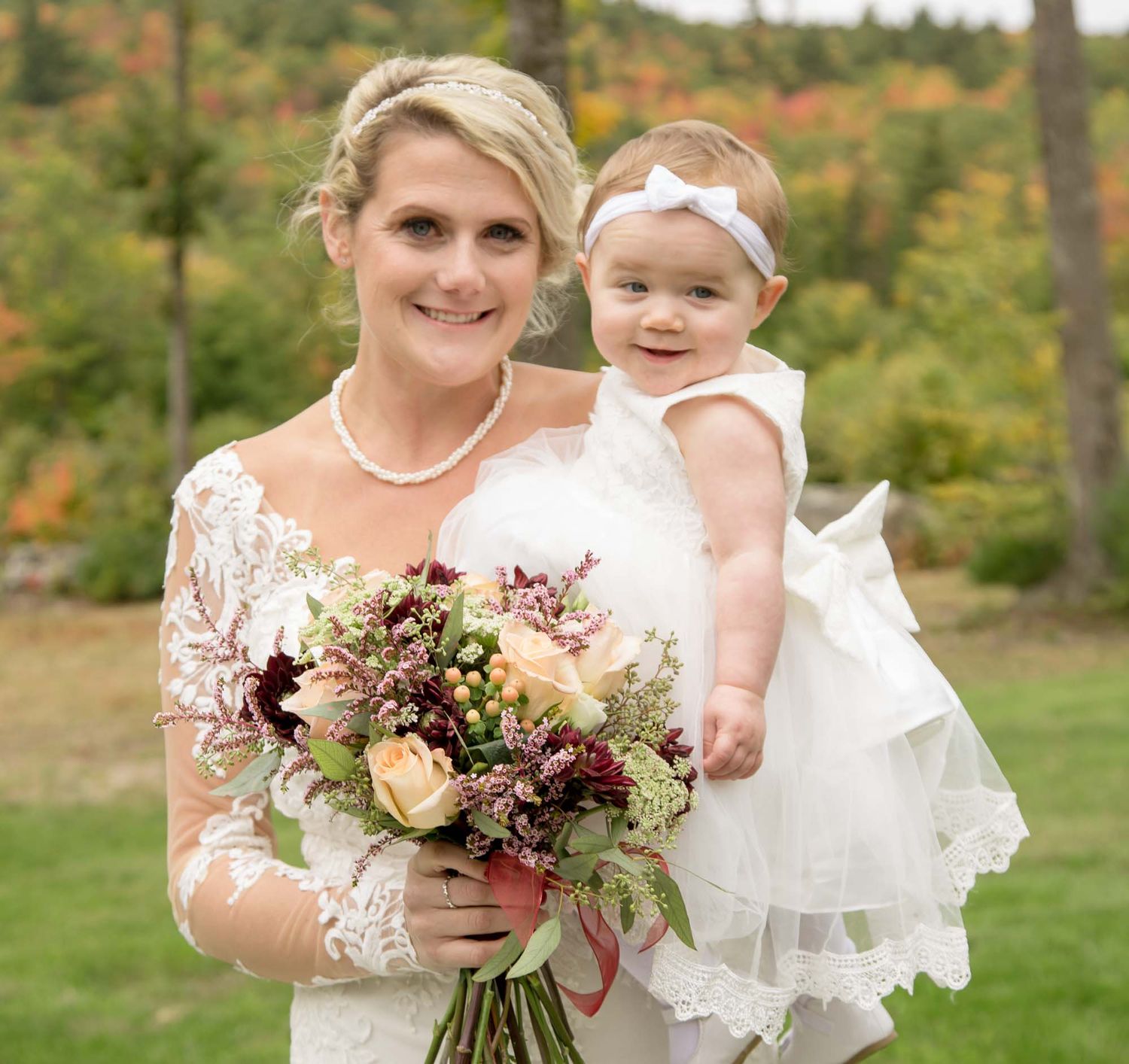 Smiling bride in lace gown holds bouquet and baby girl in white dress amid fall foliage.