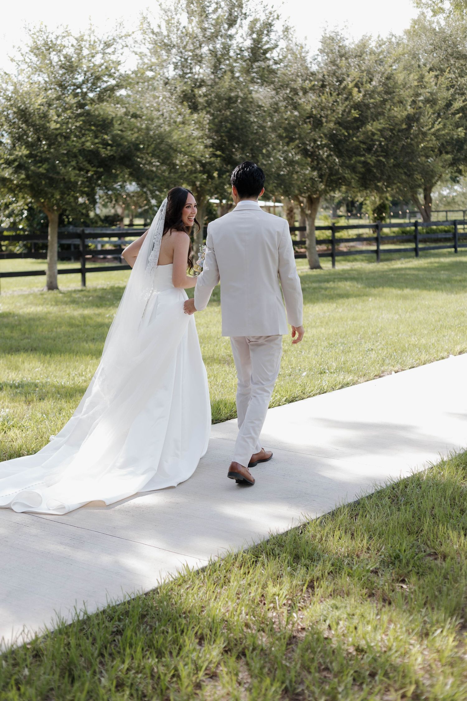 Bride and groom walking hand in hand along a white path through a lush green outdoor venue.