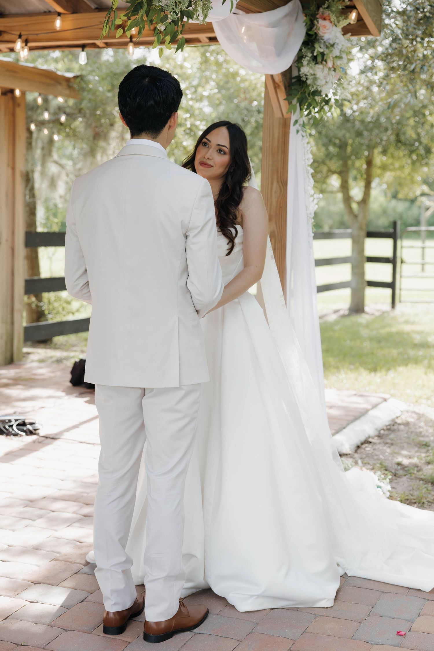 Bride and groom standing together at outdoor wedding ceremony, bride in elegant white gown with open back, groom in white suit.
