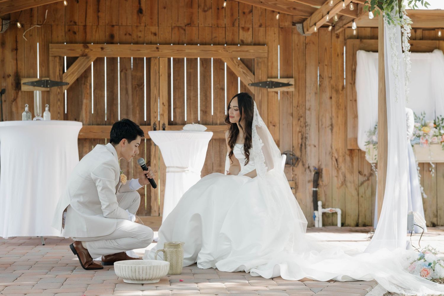 Groom kneeling before bride in white gown inside rustic barn wedding venue with draped decor.