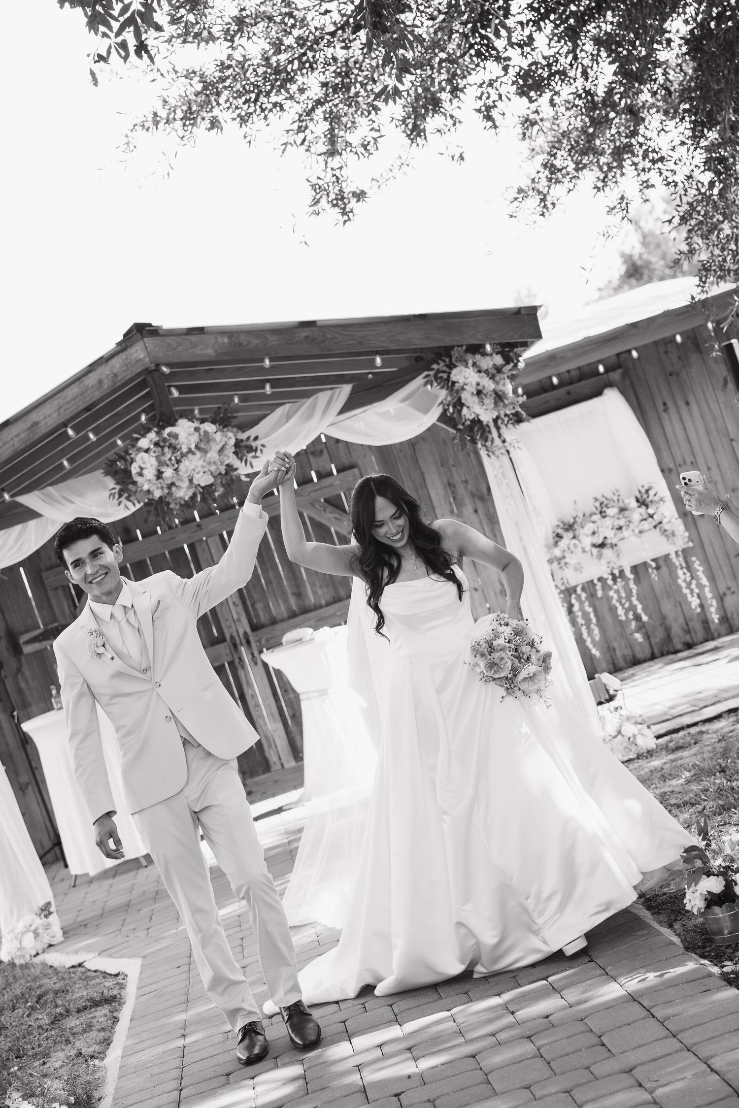 Black and white photo of joyful newlyweds celebrating after ceremony under a decorated outdoor wooden arch.