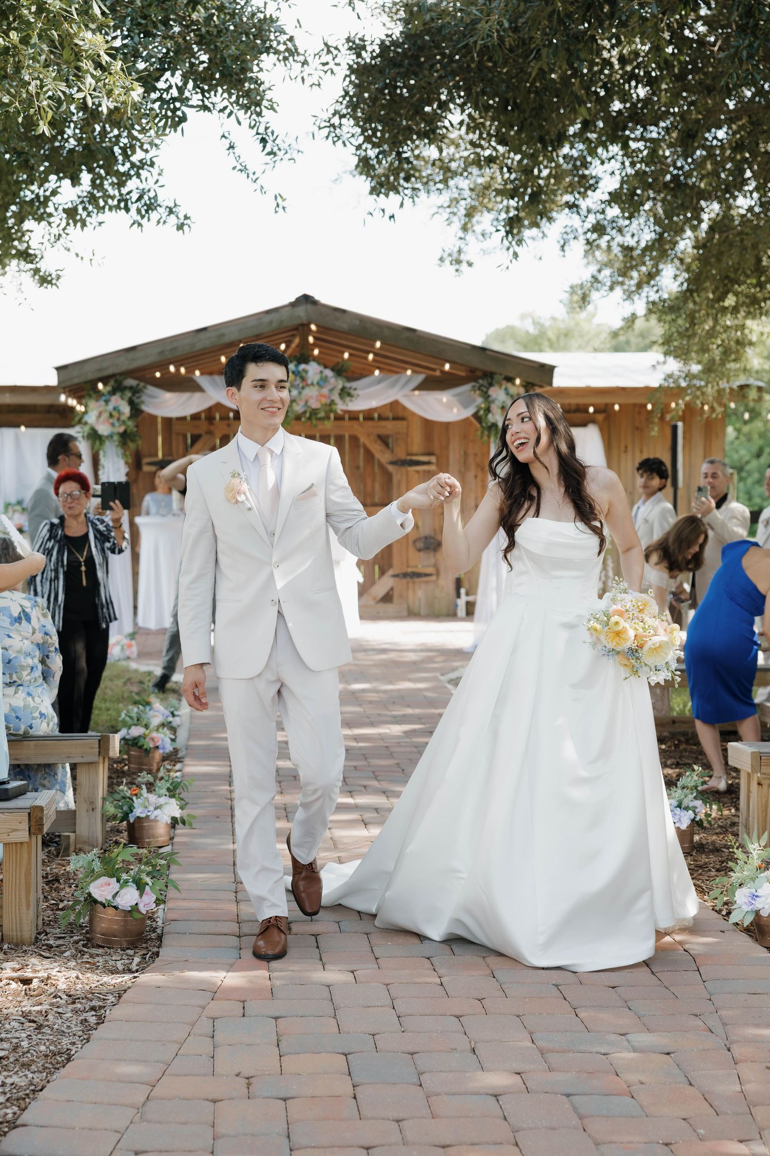 Joyful newlyweds walk hand-in-hand down brick aisle after outdoor wedding ceremony surrounded by guests.