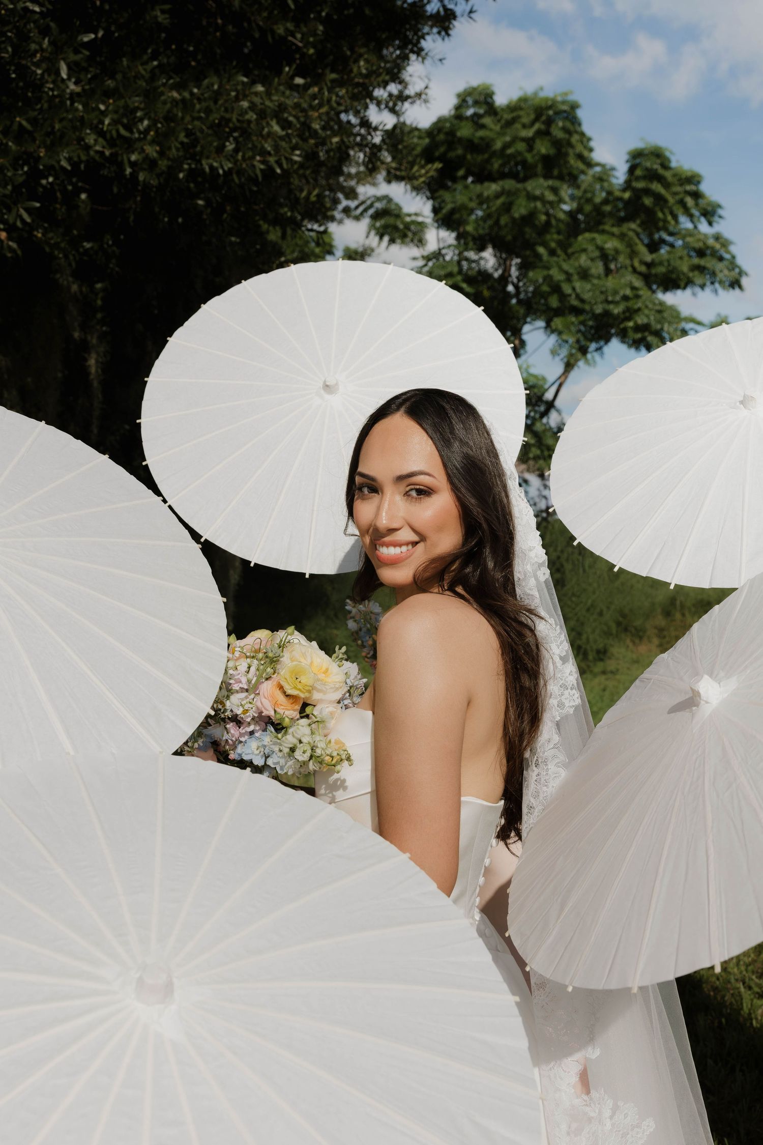 Smiling bride holds pastel bouquet surrounded by white parasols outdoors on her wedding day.