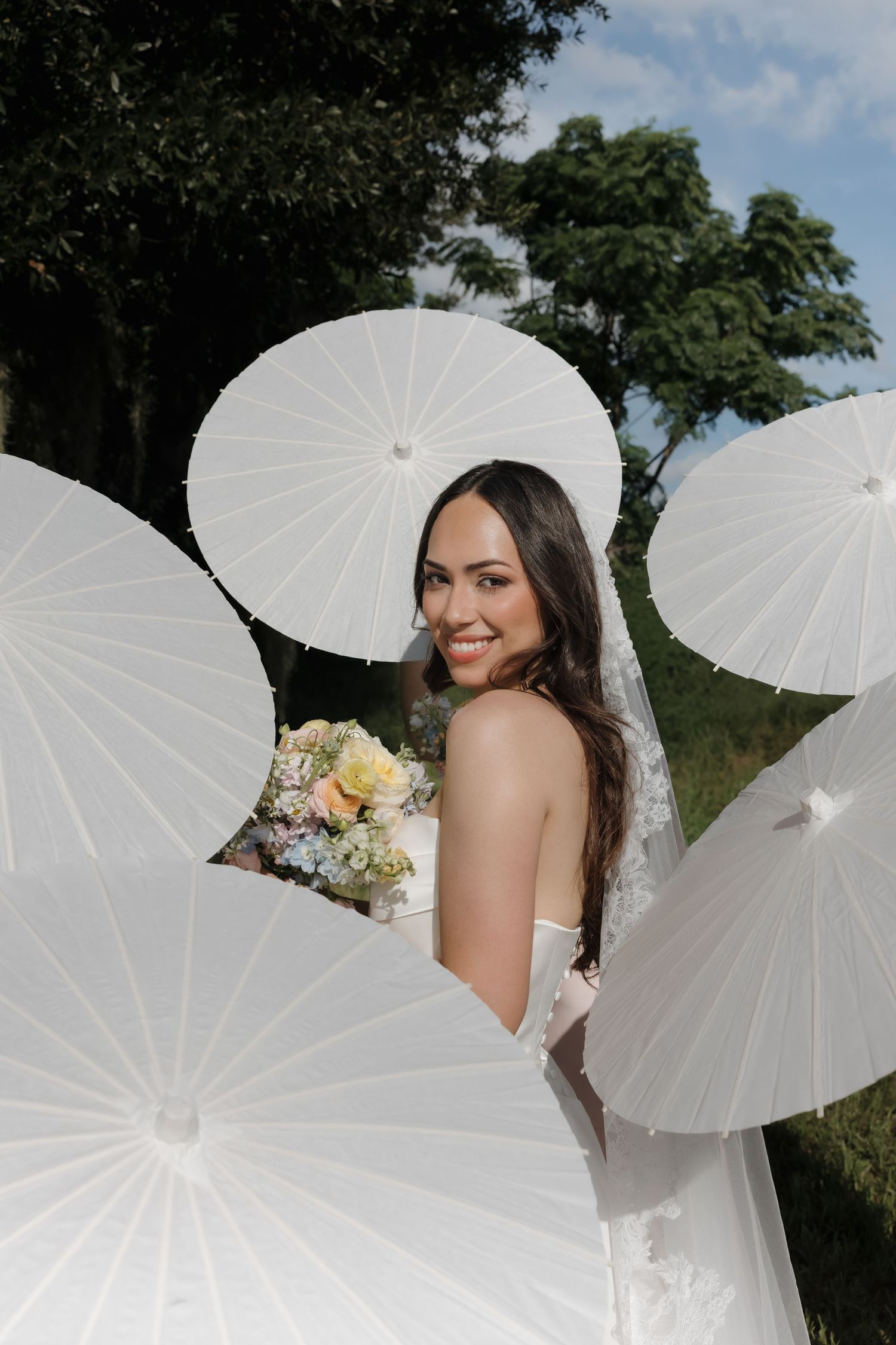 Smiling bride in white lace gown holds floral bouquet surrounded by white paper parasols outdoors.