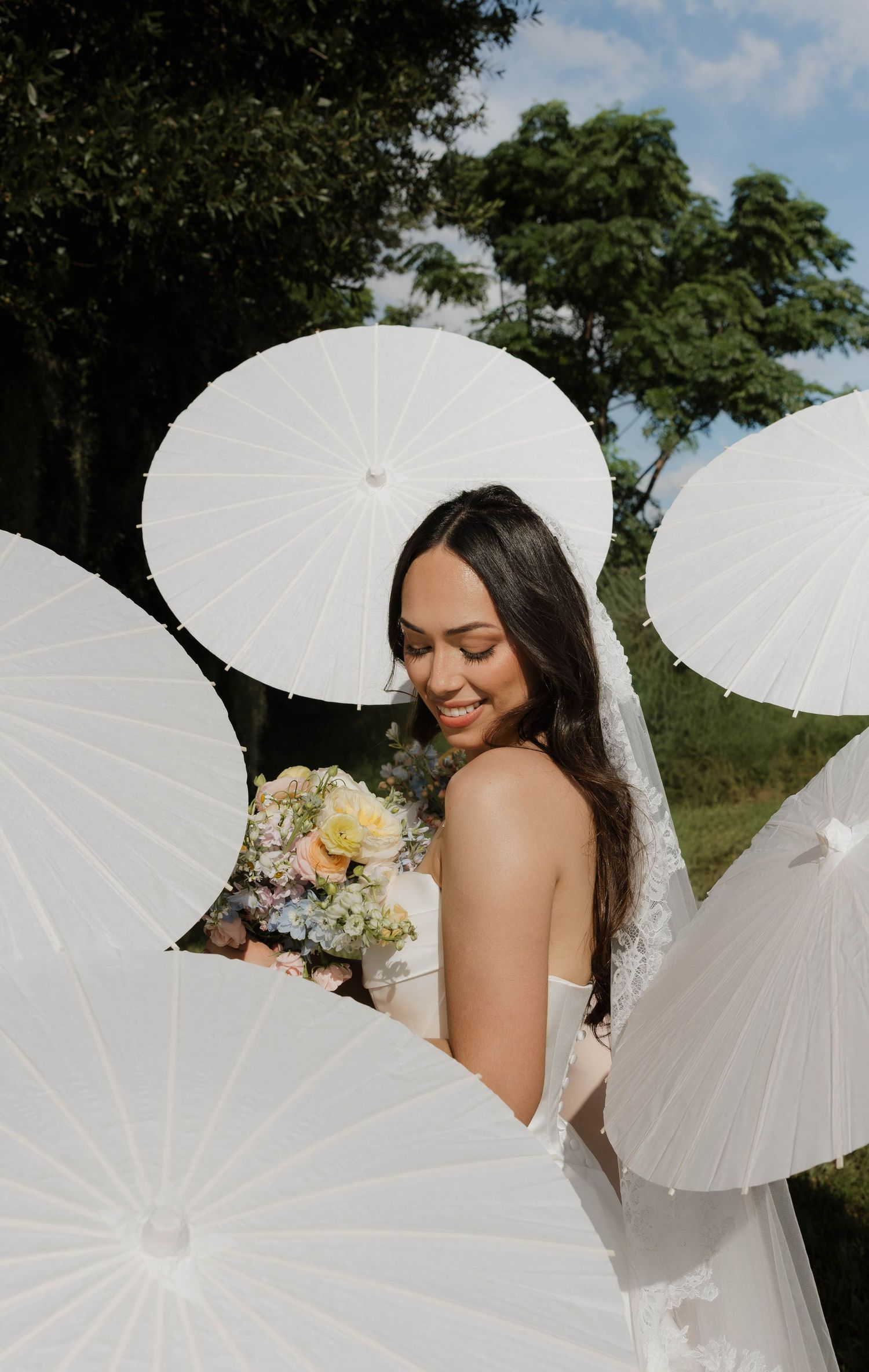 Smiling bride in white gown holds floral bouquet surrounded by white parasols outdoors.