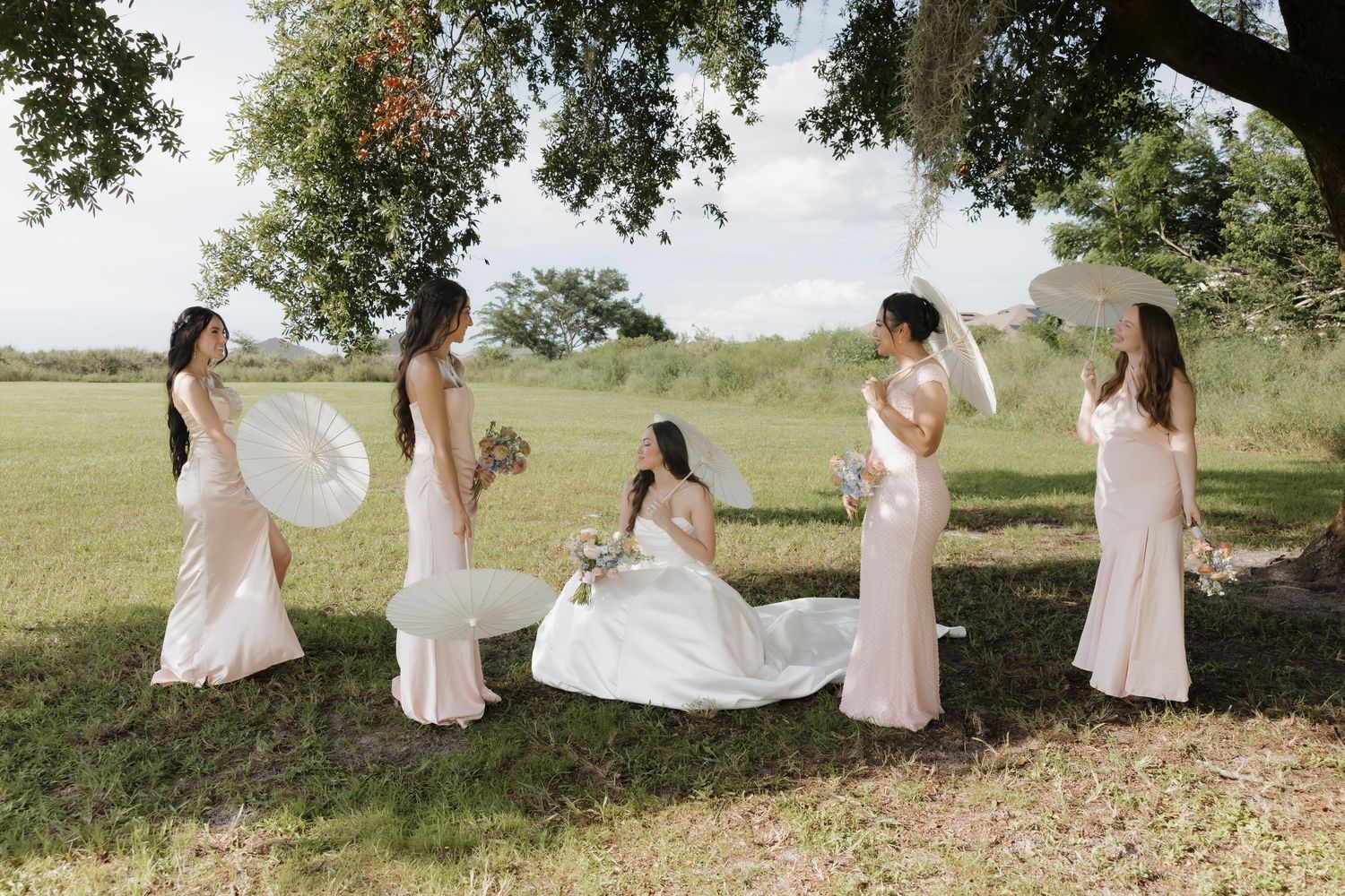 Bride in white gown sits outdoors with four bridesmaids in blush dresses holding parasols and bouquets under a shady tree.