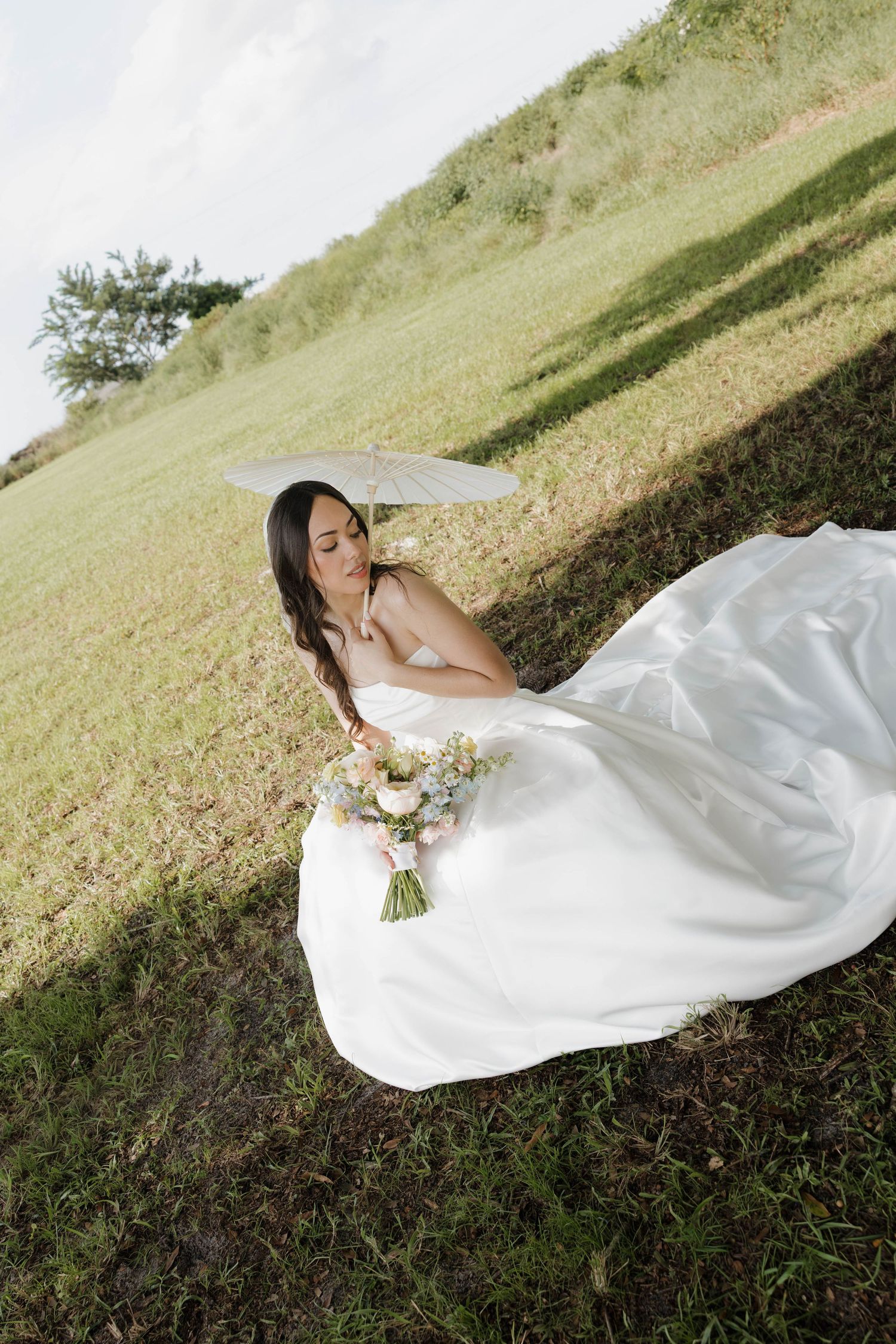 Bride in flowing white strapless gown sits on green hillside holding a pastel bouquet under a white parasol.