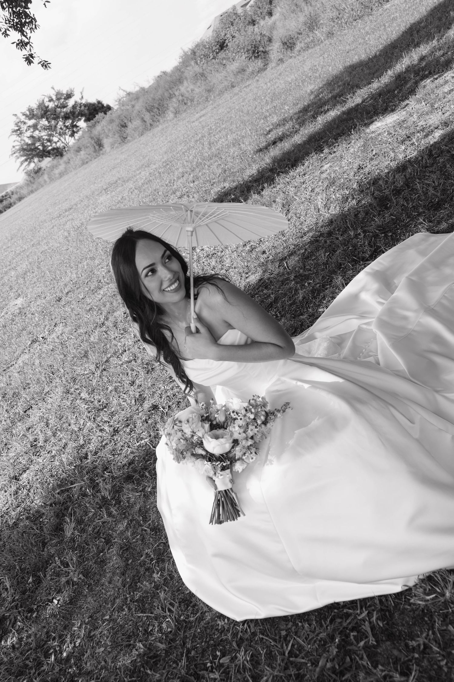 Black and white photo of bride in flowing gown holding parasol and bouquet in open field.