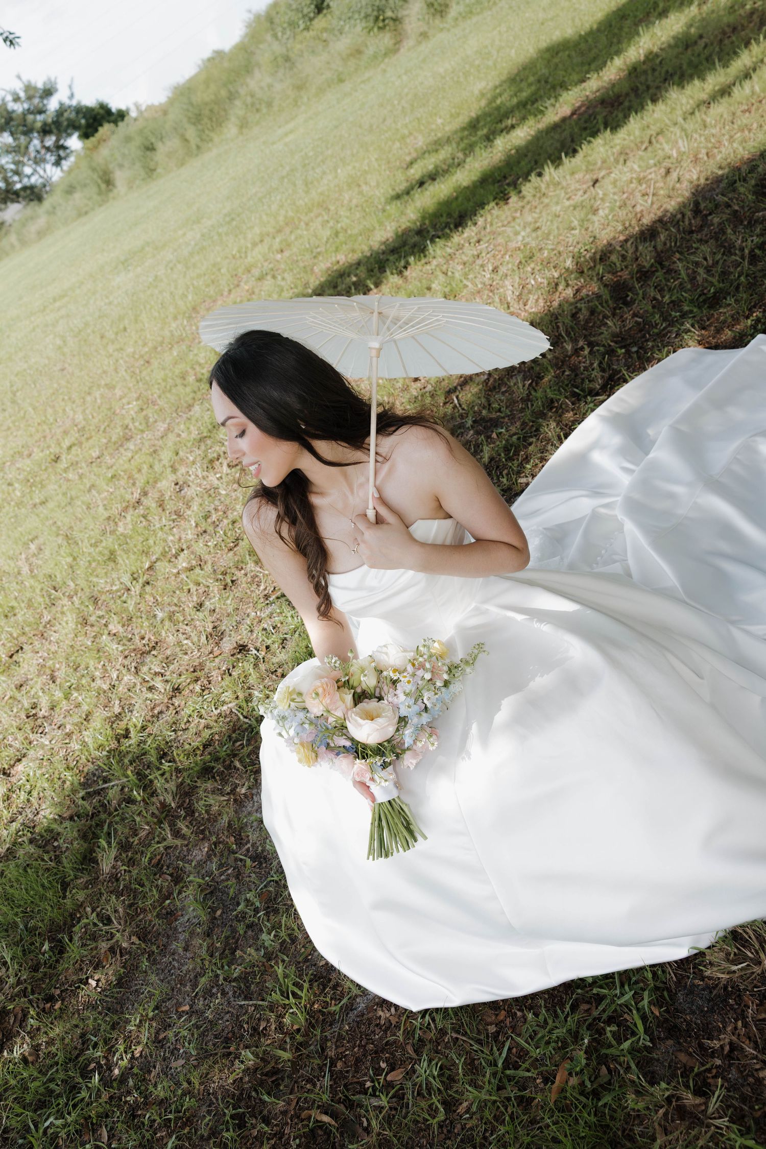 Aerial view of bride in white gown holding parasol and pastel bouquet in grassy outdoor setting.
