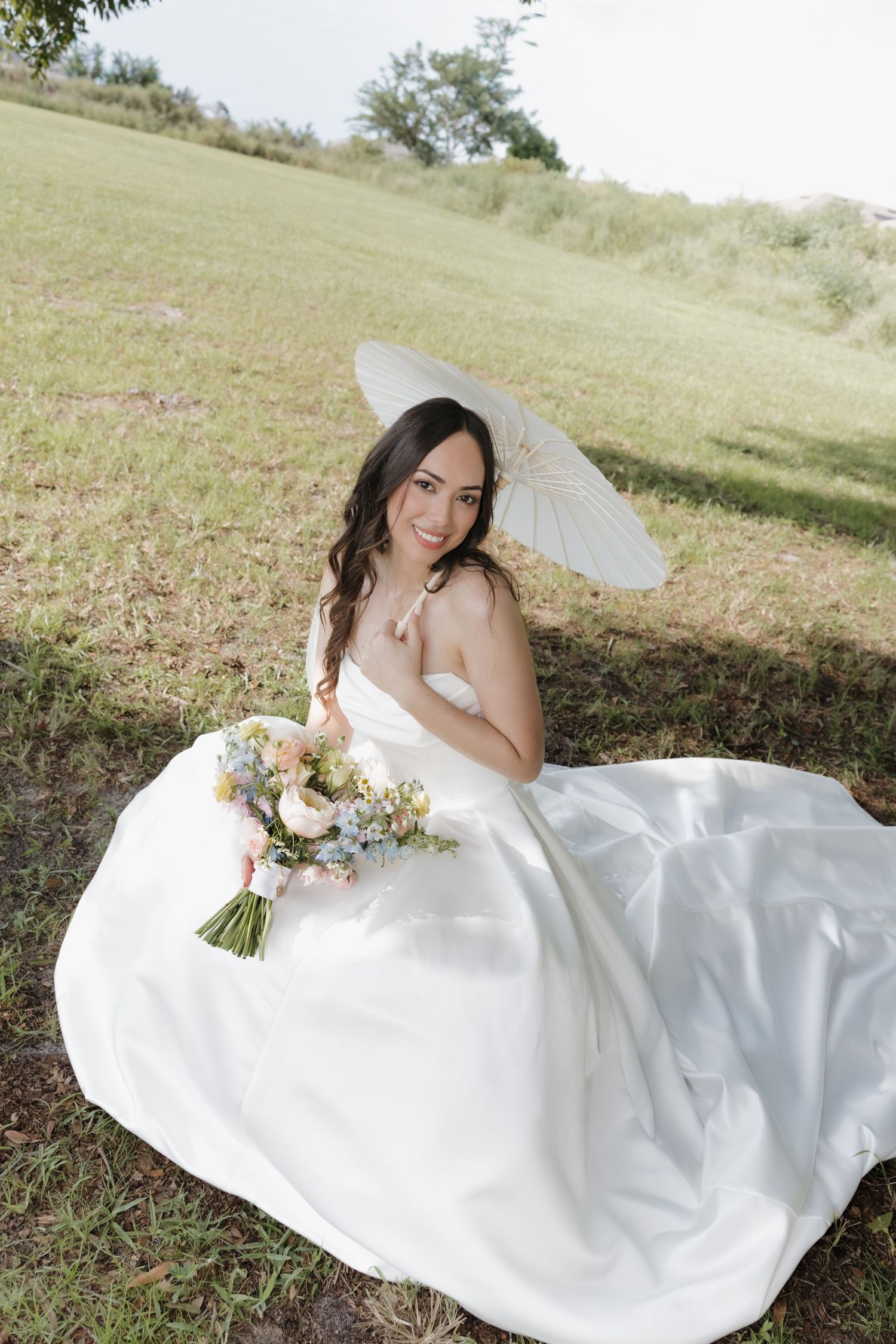 Smiling bride in white gown sits on grass holding bouquet and parasol.