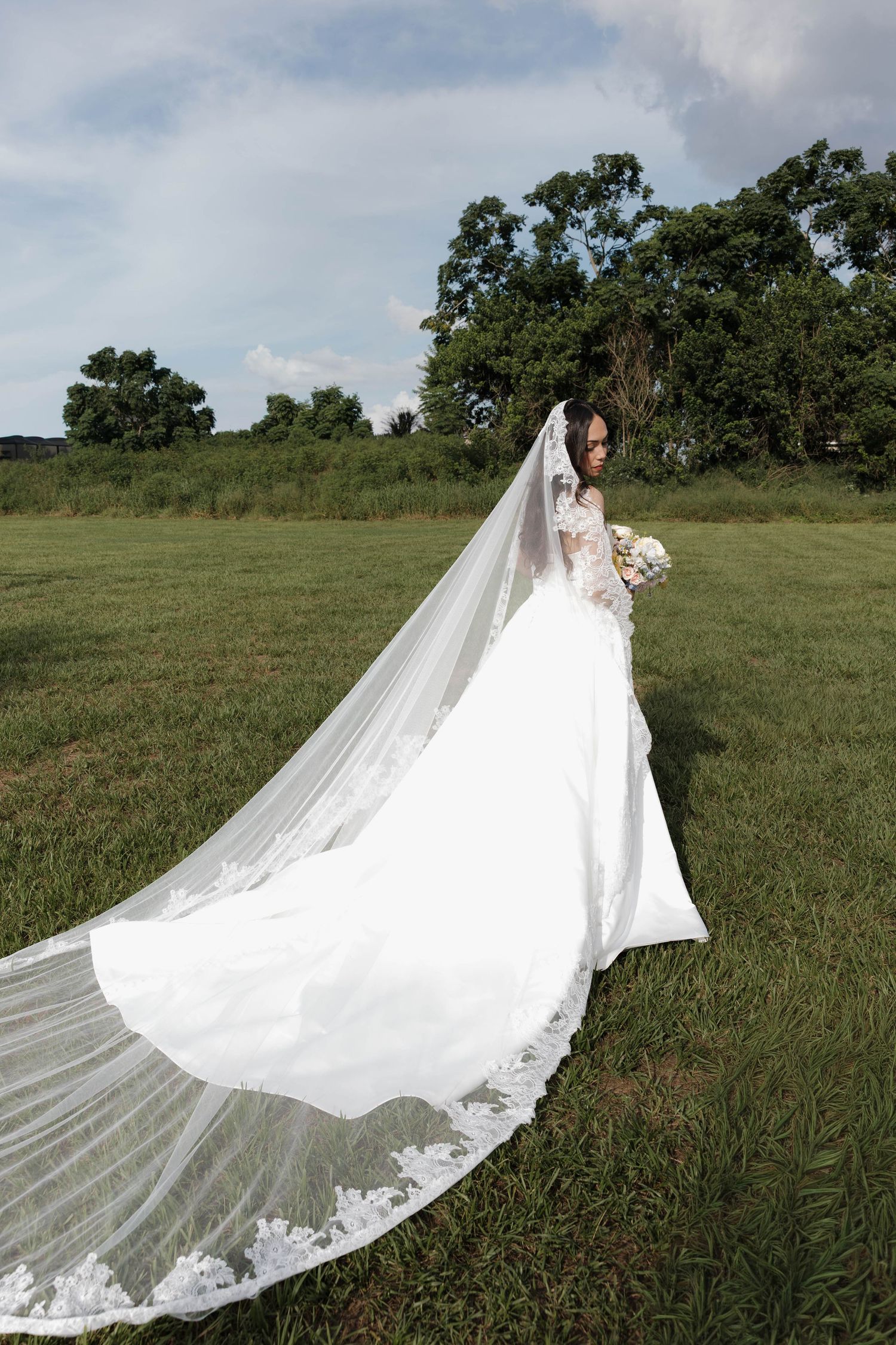 Bride in a flowing white gown with long lace veil stands in a green field holding a bouquet.