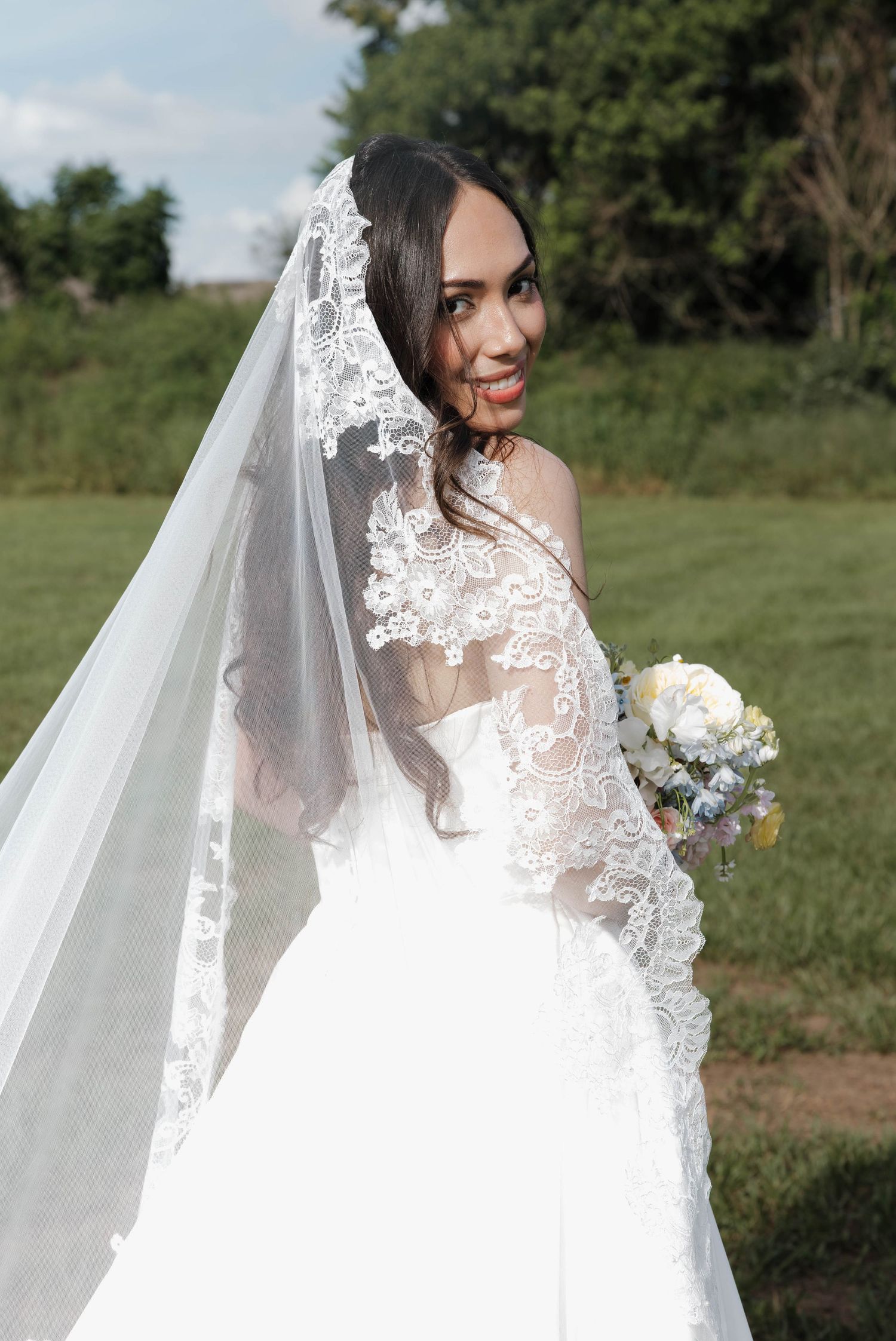 Smiling bride in a white lace wedding gown and long veil holding a bouquet outdoors on her wedding day.