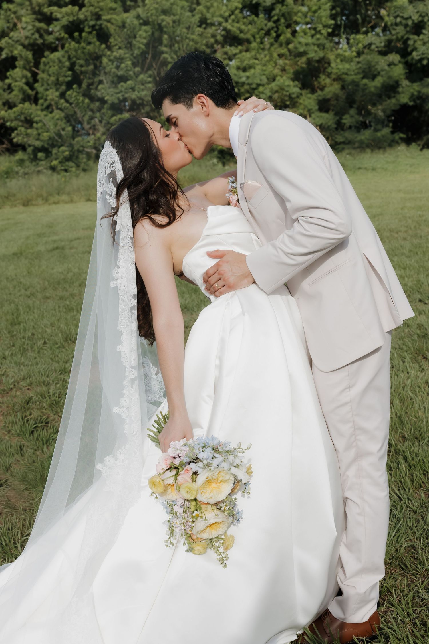 Bride and groom share a romantic kiss outdoors, she holds a pastel bouquet in a white strapless gown with lace veil.