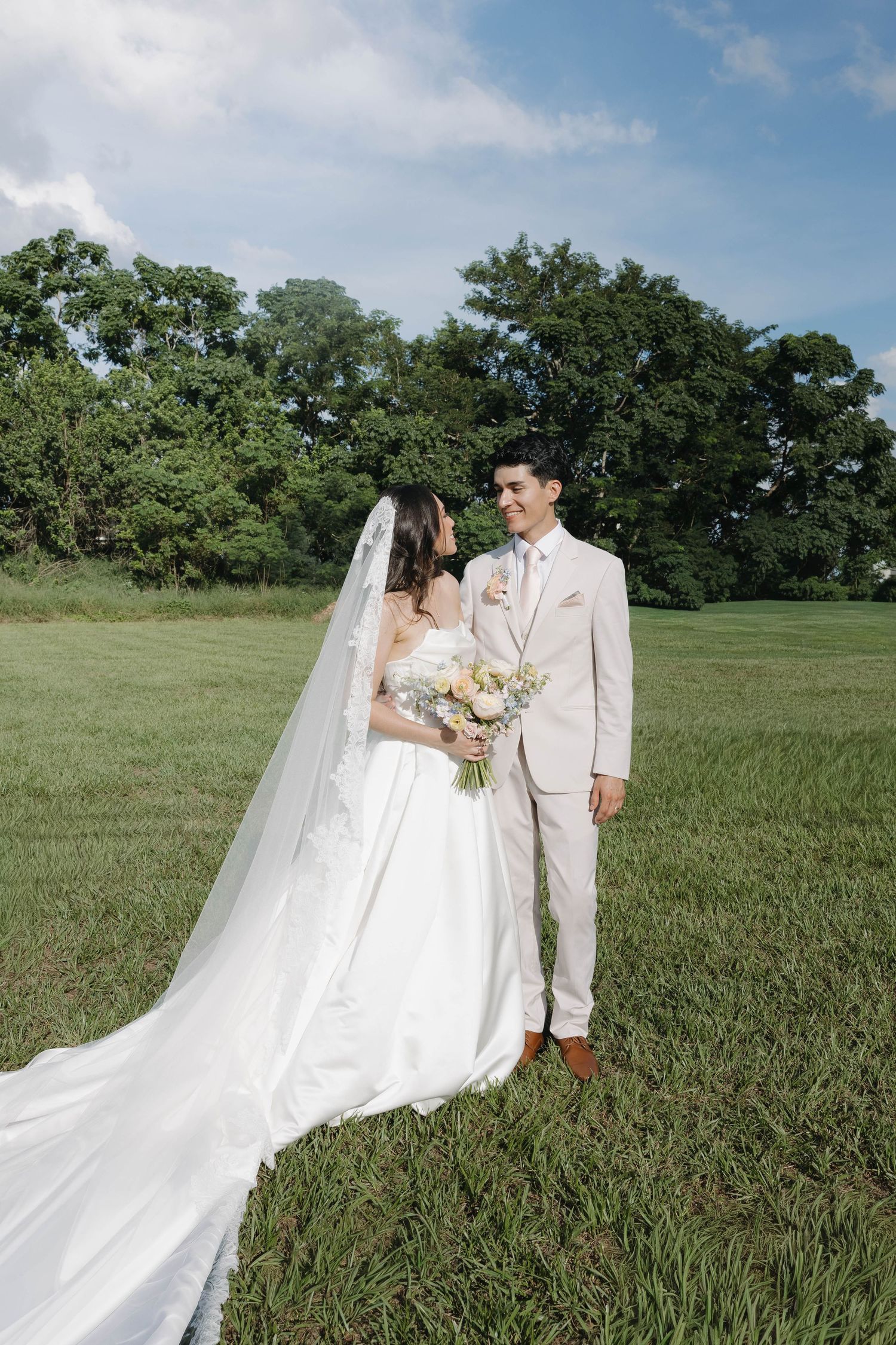 Bride and groom facing each other smiling in a green meadow under a partly cloudy sky.