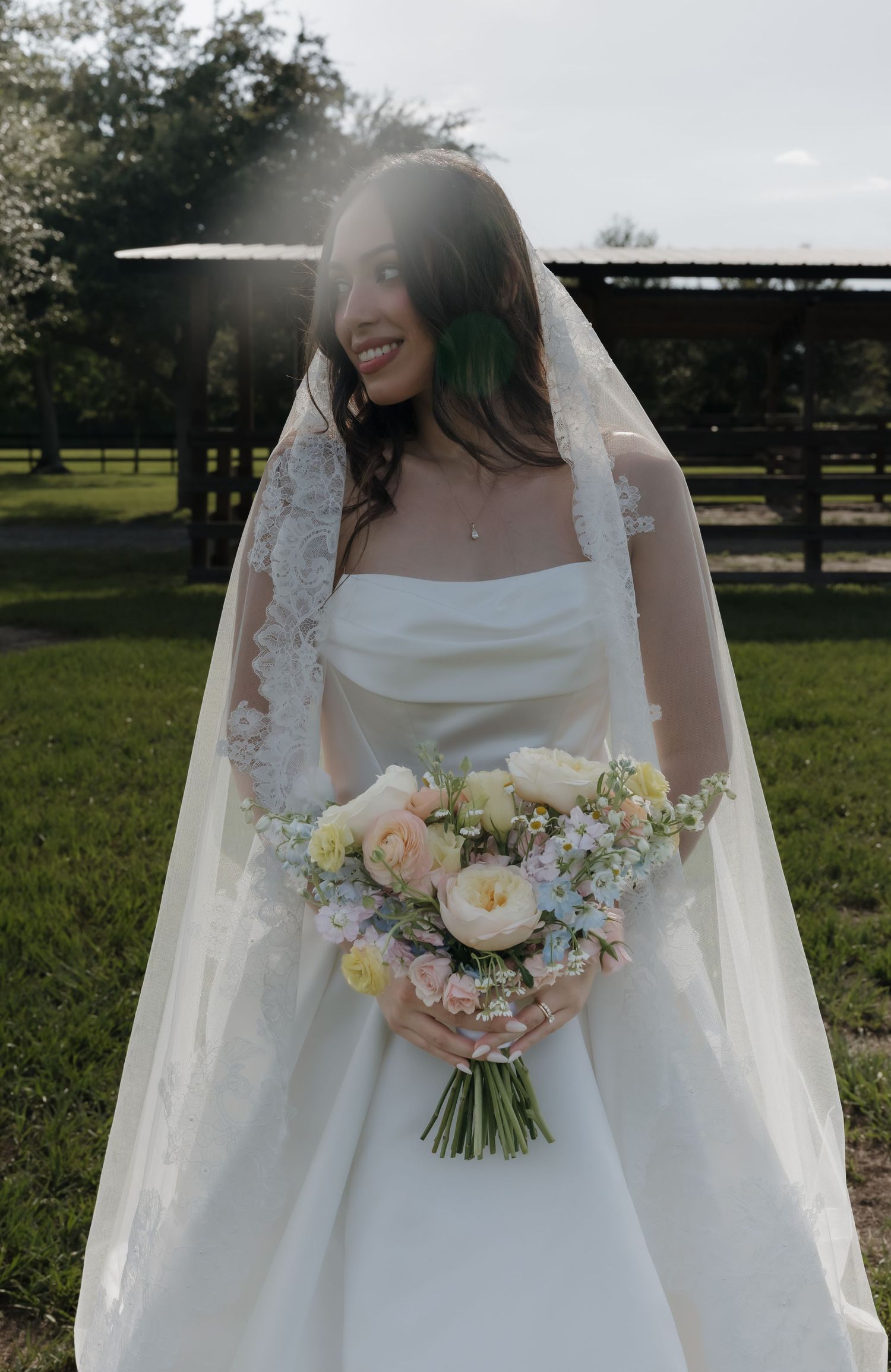Smiling bride in white gown and lace veil holds pastel bouquet outdoors on green lawn with sunlight behind her.