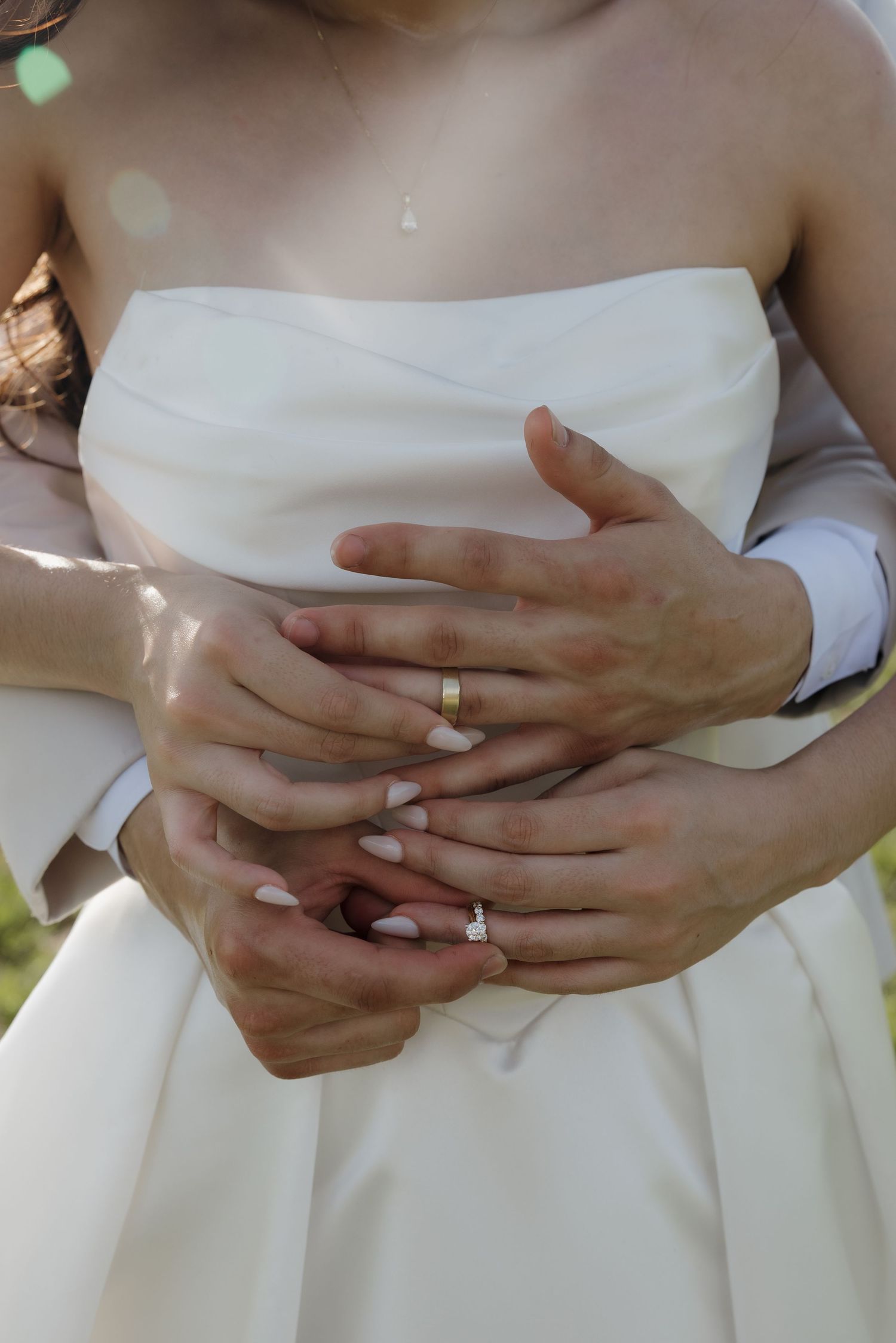 Bride and groom holding hands showing wedding rings, woman in white strapless gown embraced from behind by partner.