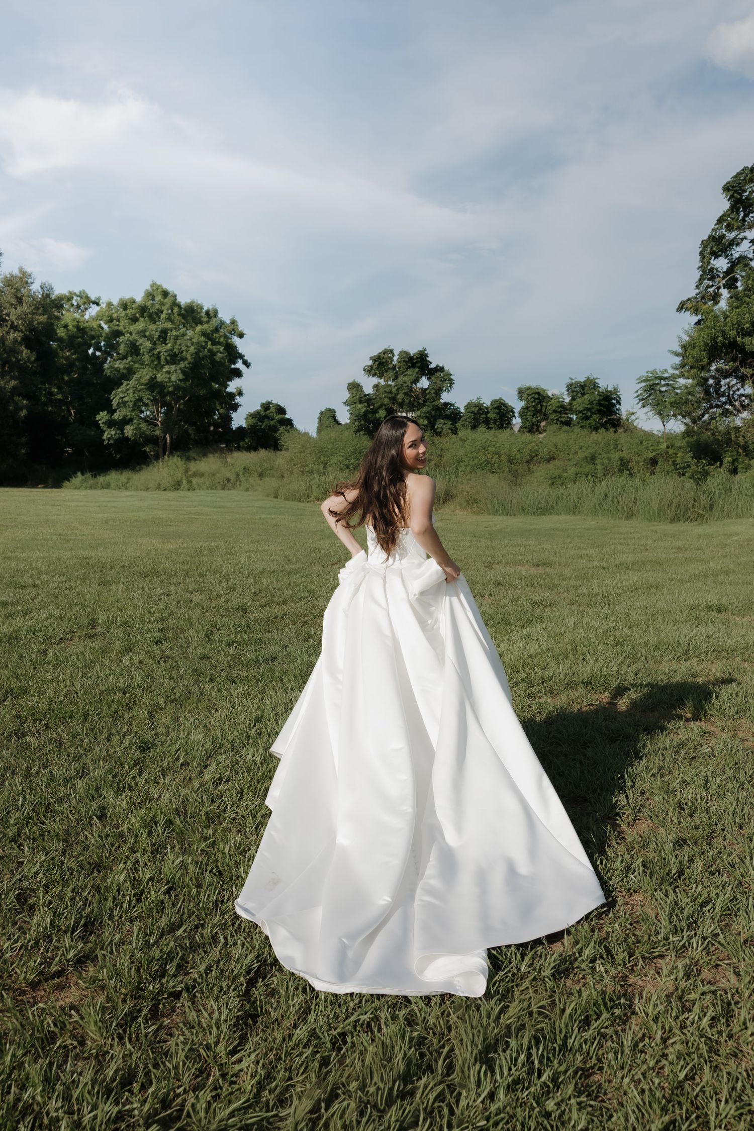 Bride in a flowing white ball gown twirling in a lush green field with trees in the background.