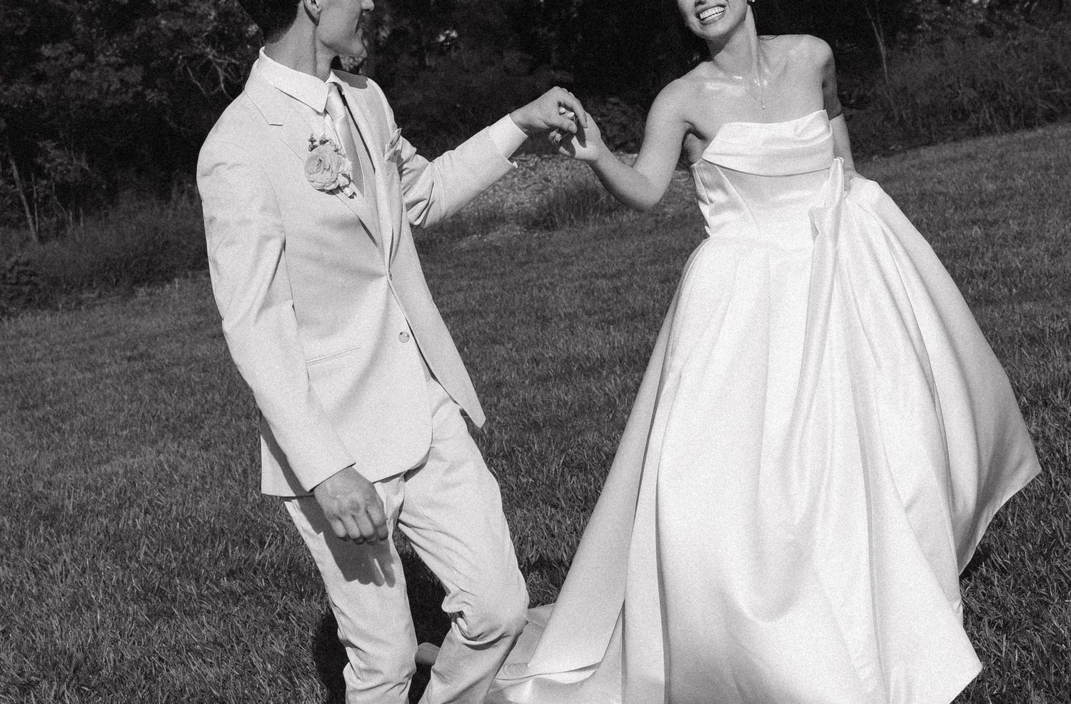 Black and white photo of a couple in wedding attire dancing outdoors, bride in strapless gown, groom in white suit.