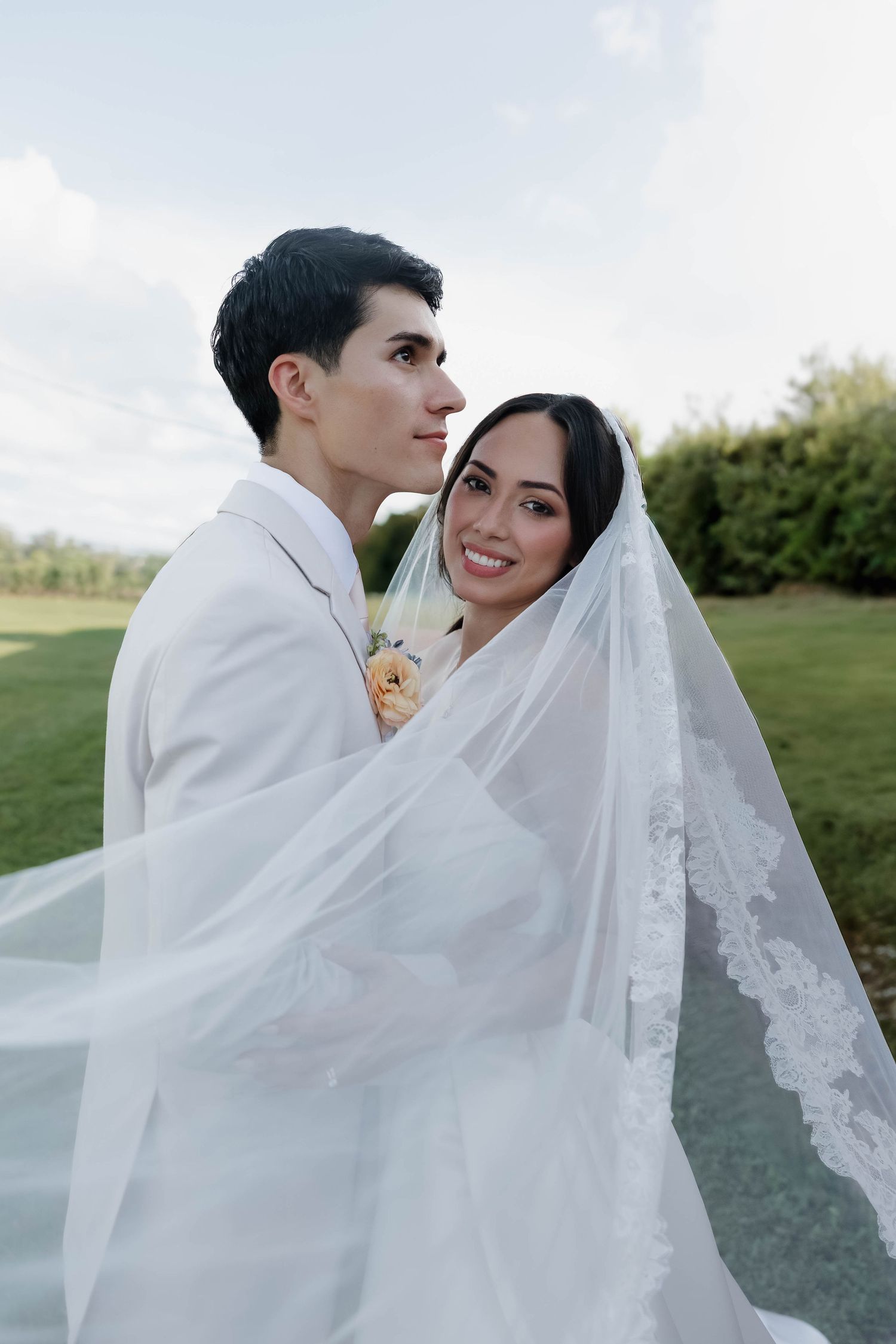 Bride and groom wrapped in a flowing lace veil outdoors on their wedding day, smiling in a lush green garden.