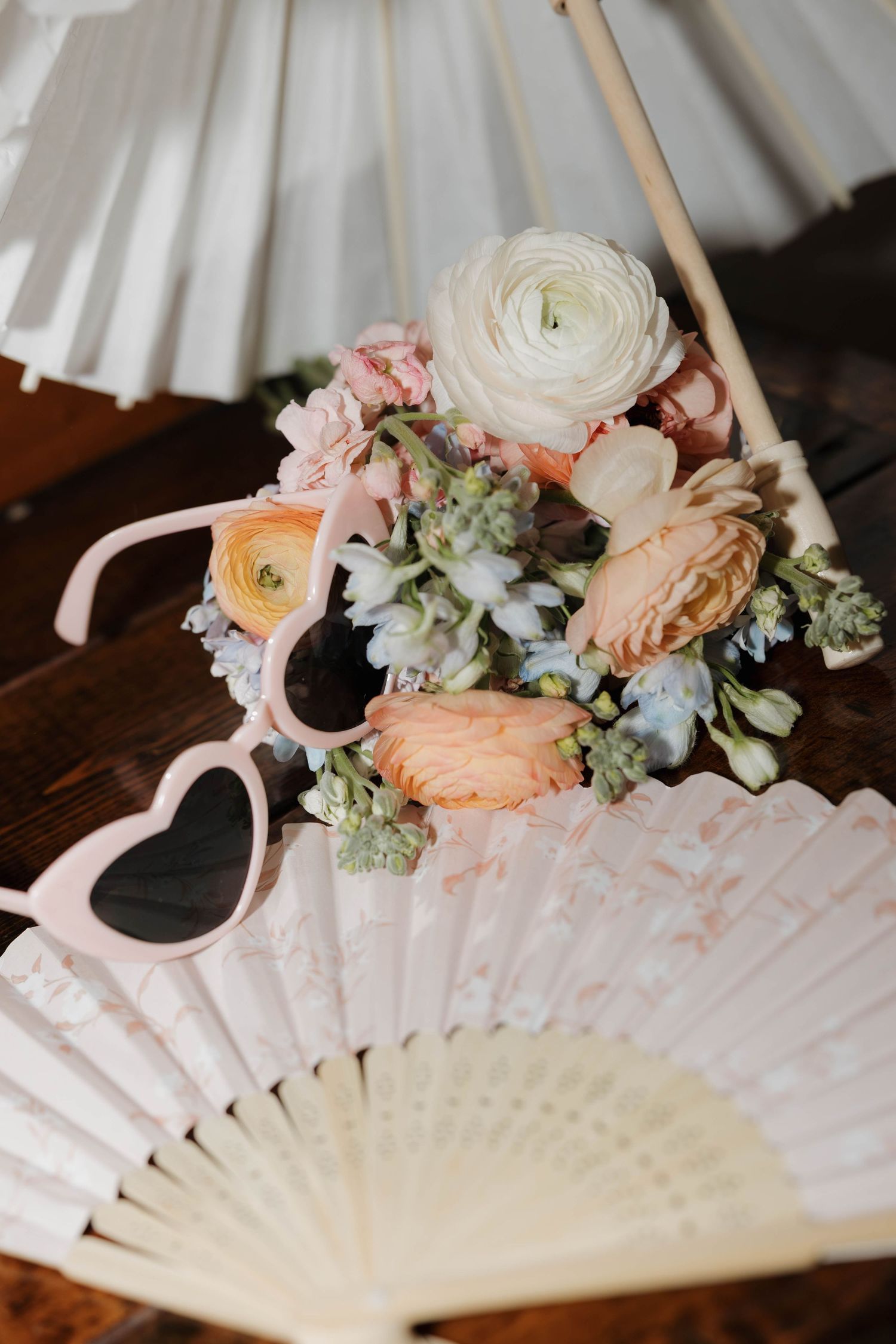 Wedding accessories flat lay featuring pastel flowers, heart-shaped sunglasses, and decorative hand fans on wood.