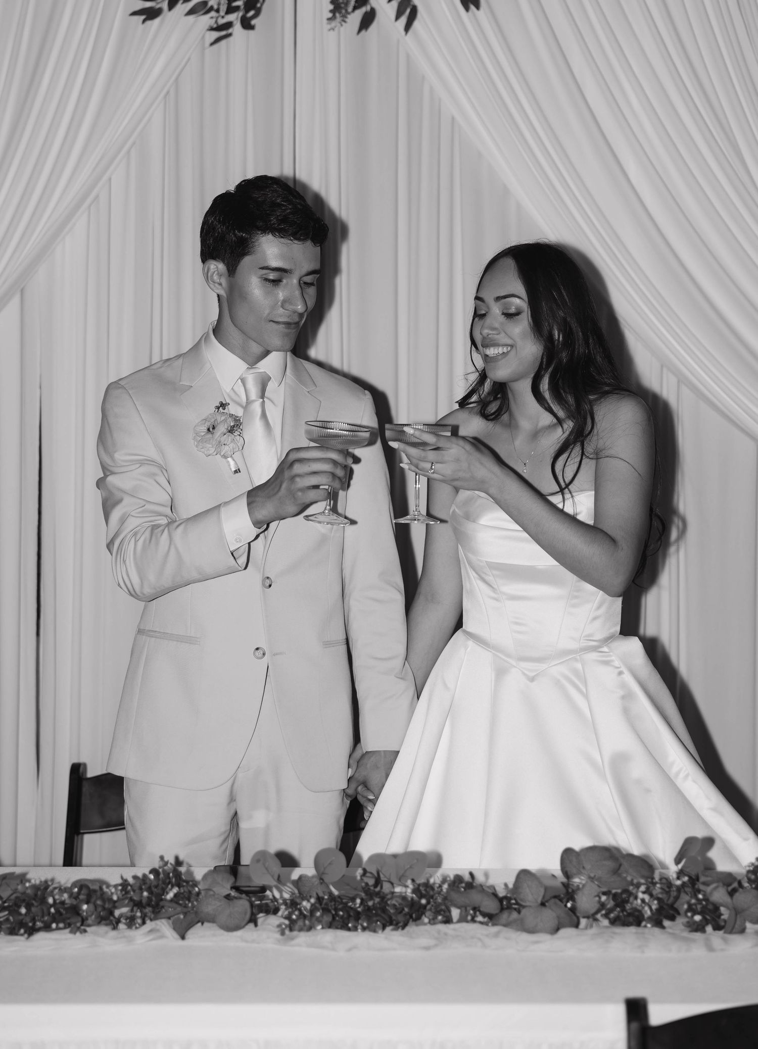 Black and white photo of a bride and groom toasting with champagne glasses at their wedding reception.