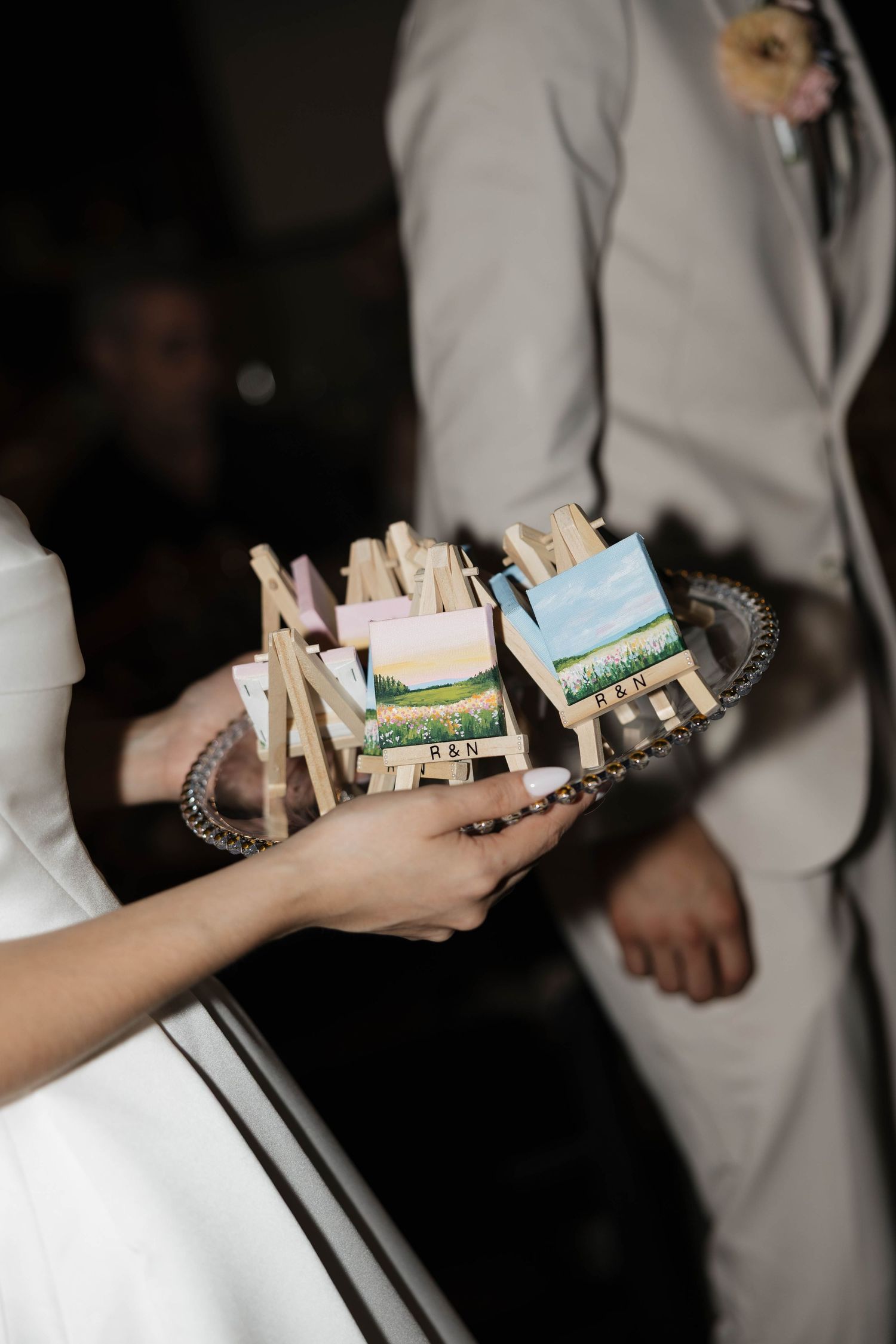 Bride holds a silver tray displaying miniature easels with tiny landscape paintings as wedding favors.