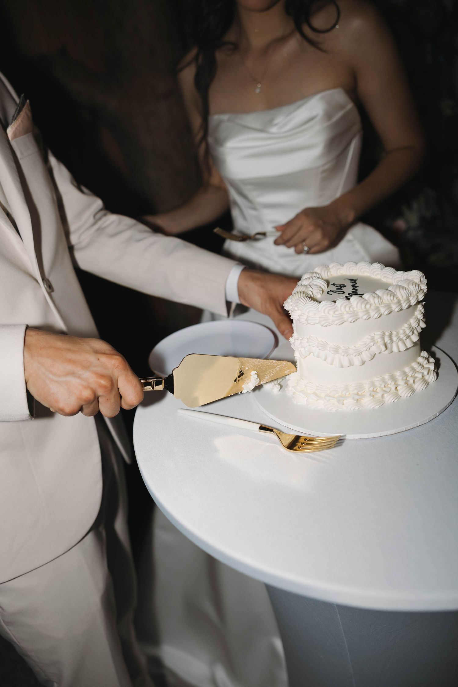 Bride and groom cutting a two-tier white wedding cake with gold cake server on a round white table.