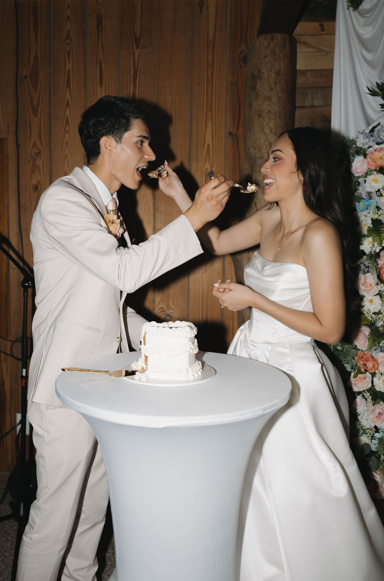 Bride and groom feeding each other wedding cake at rustic barn reception with floral arrangements.