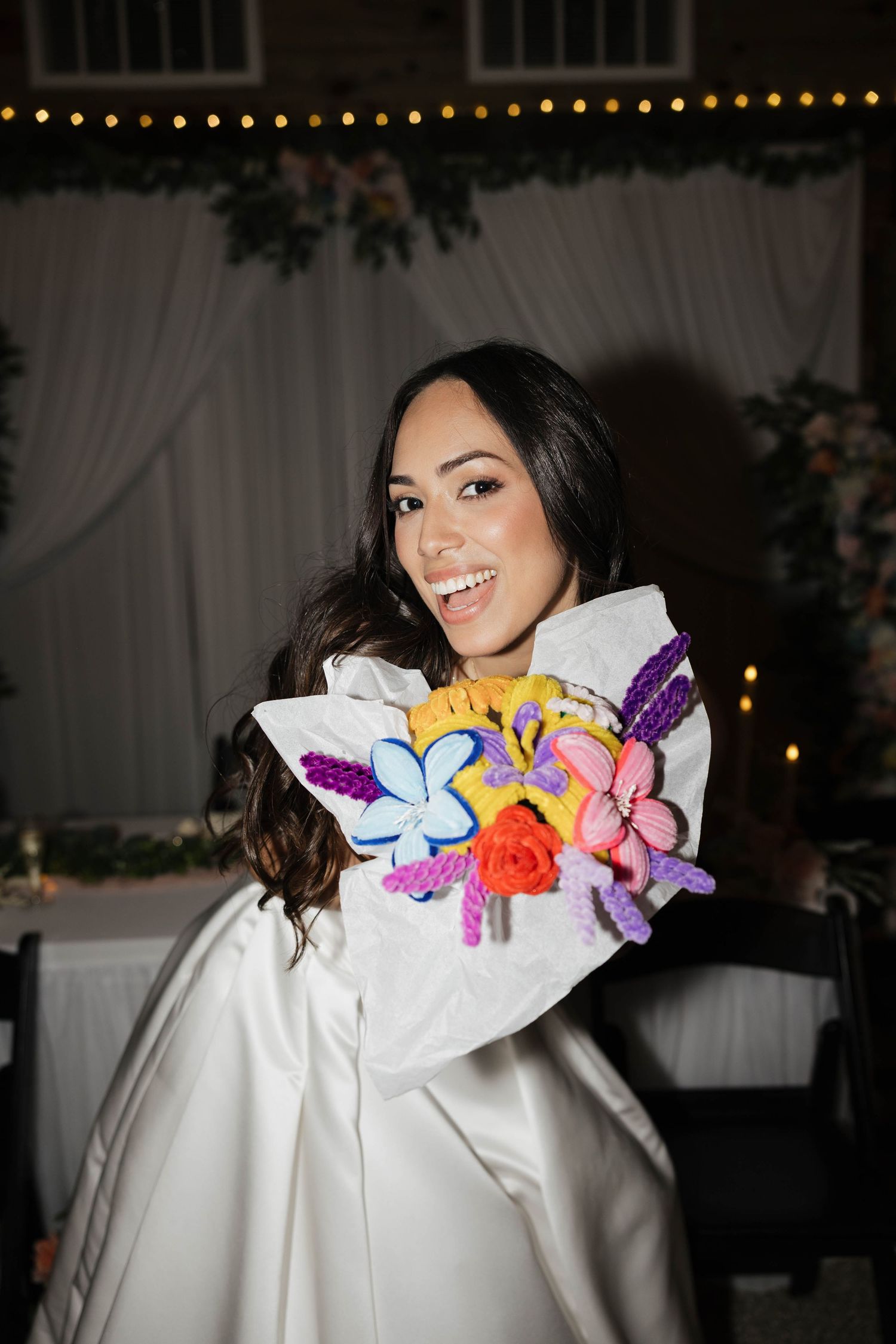 Smiling bride in white gown holding colorful embroidered floral bouquet at outdoor wedding reception.
