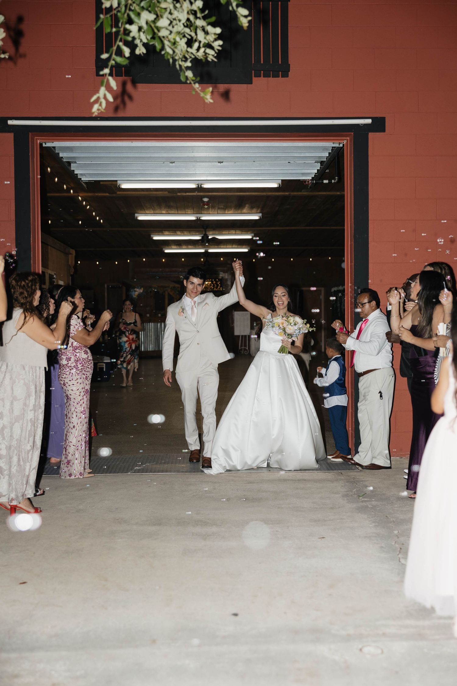 Bride and groom exit wedding reception through garage door entrance, guests cheering and blowing bubbles in celebration.