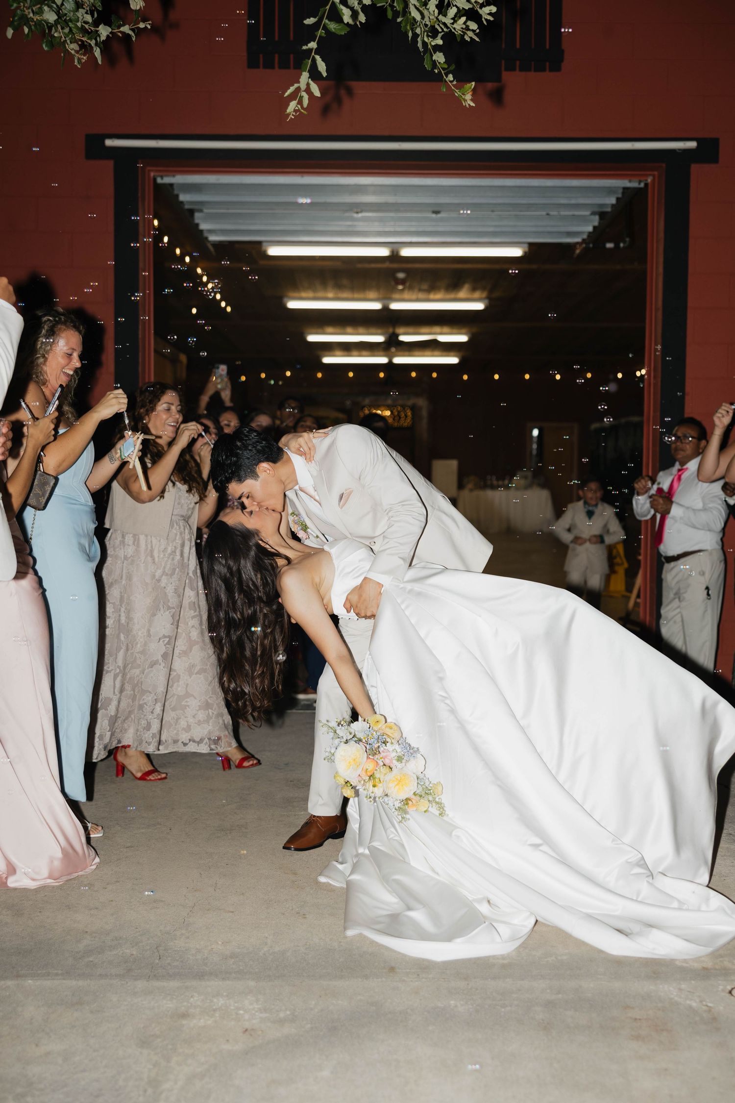 Bride and groom share a romantic dip kiss at wedding exit surrounded by cheering guests blowing bubbles.