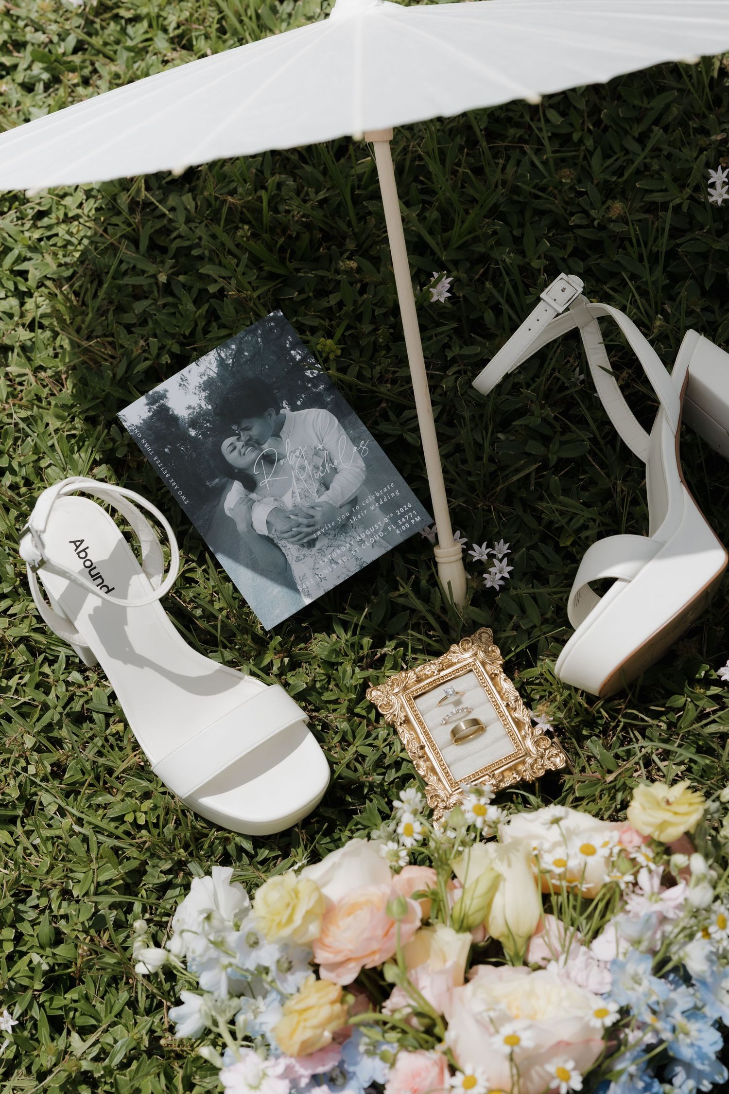 White bridal heels, wedding rings in gold frame, floral bouquet, photo book, and parasol arranged on green grass.