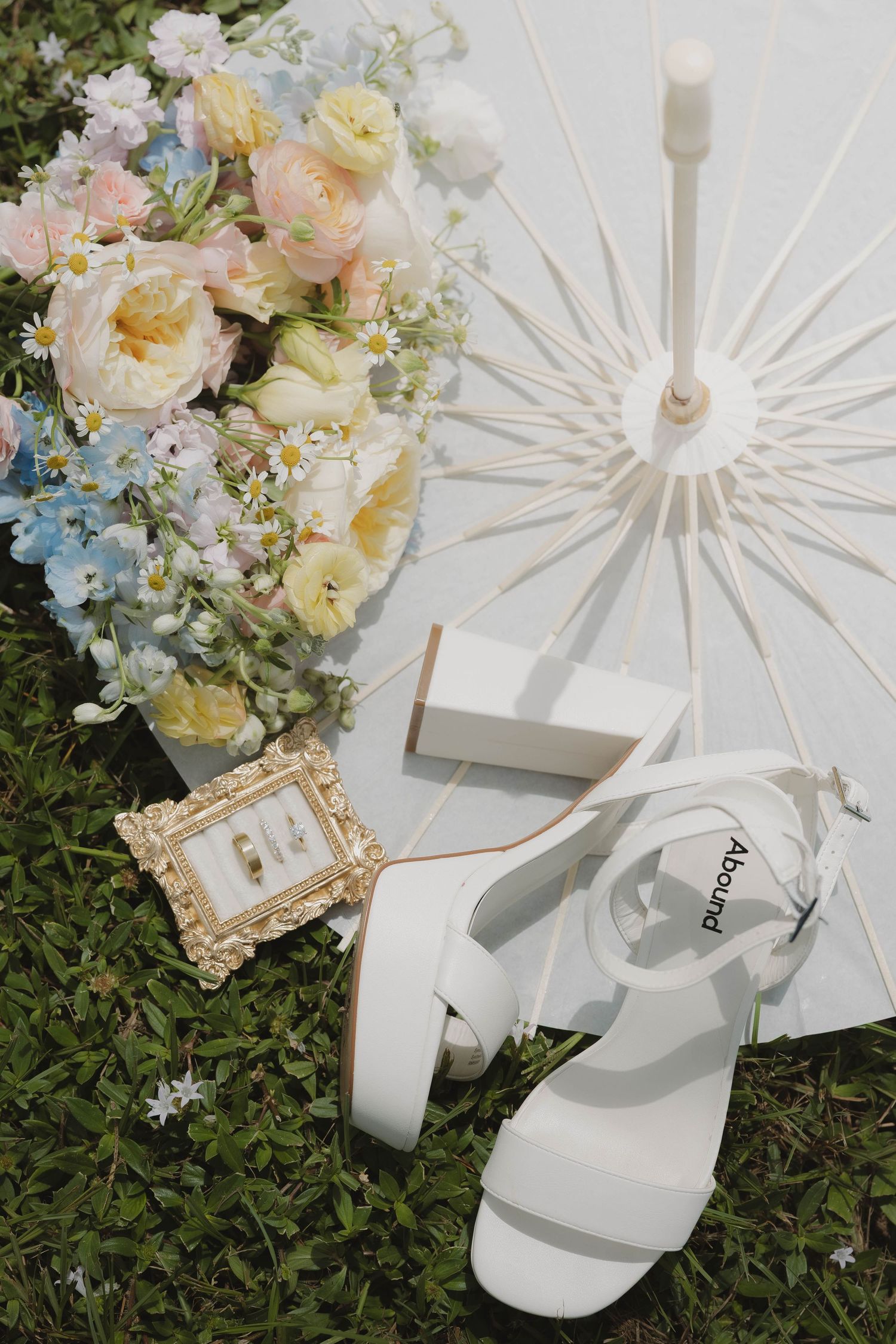 Wedding flat lay with bouquet, white heels, parasol, and rings on grass.