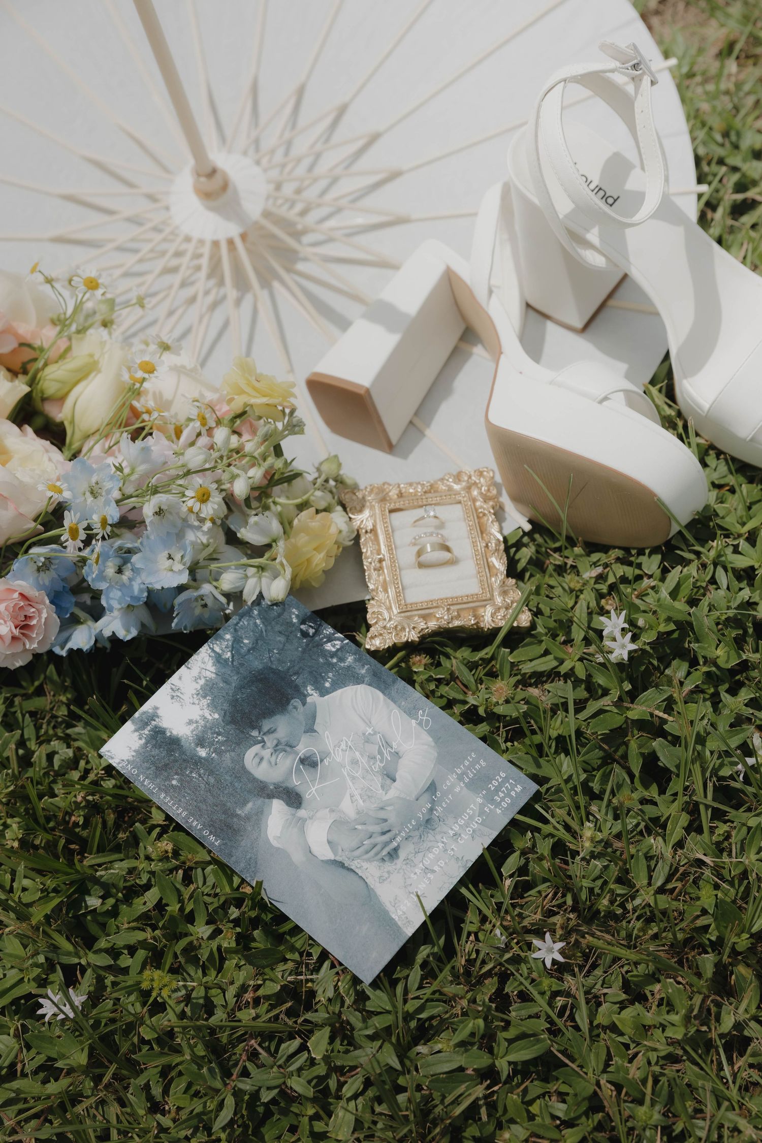 Wedding flat lay with white heels, floral bouquet, parasol, ring box, and vintage photo on grass.
