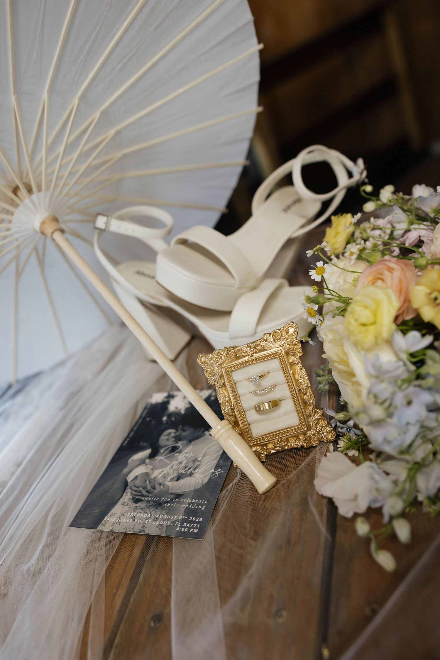Wedding flat lay with white parasol, heels, gold framed rings, bouquet, and invitation on wood.