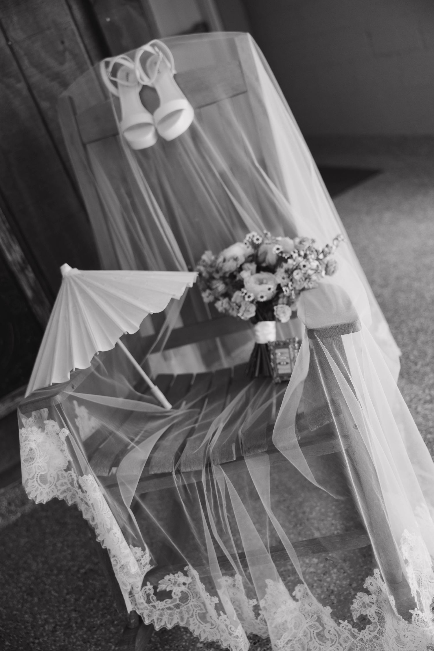 Black and white photo of bridal veil, bouquet, parasol, and white heels arranged on a wooden bench.