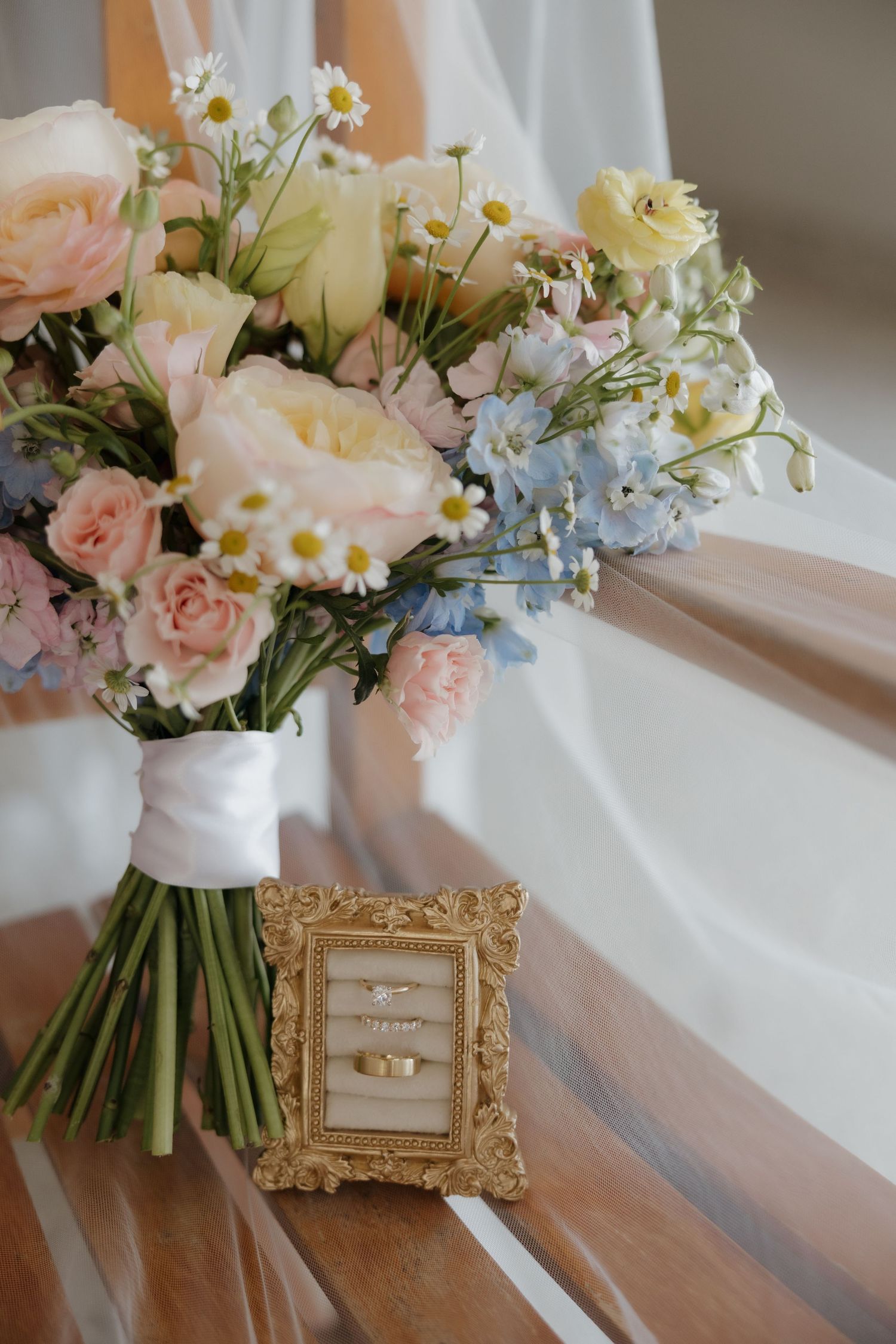 Elegant bridal bouquet of pink roses, blue hydrangeas, and daisies beside a gold frame with wedding rings on wood.